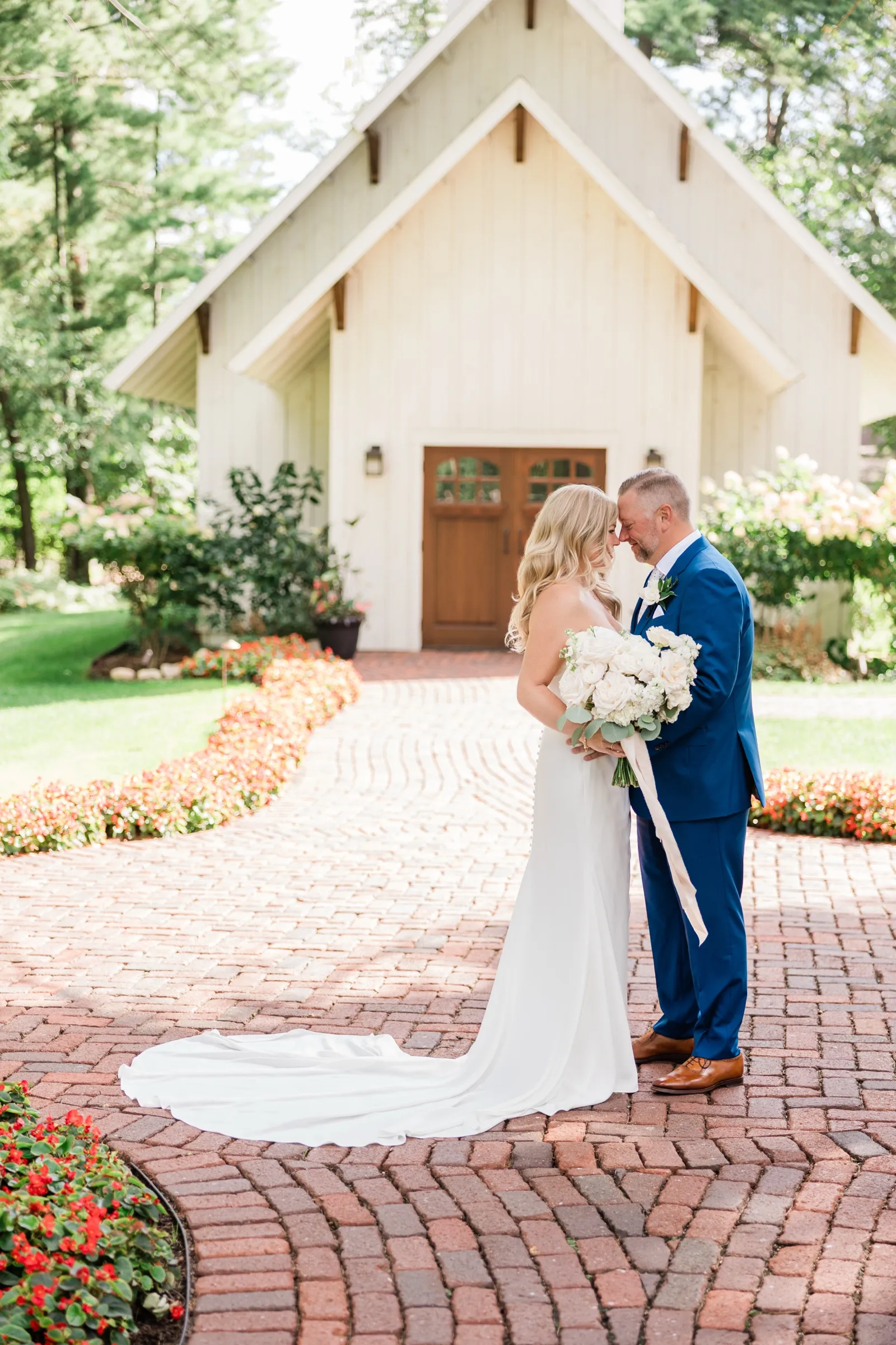 The bride and groom stand forehead to forehead in front of the white chapel doors, her train sweeping across the curved brick courtyard. — Tim Larsen Photography, Brainerd Lakes MN