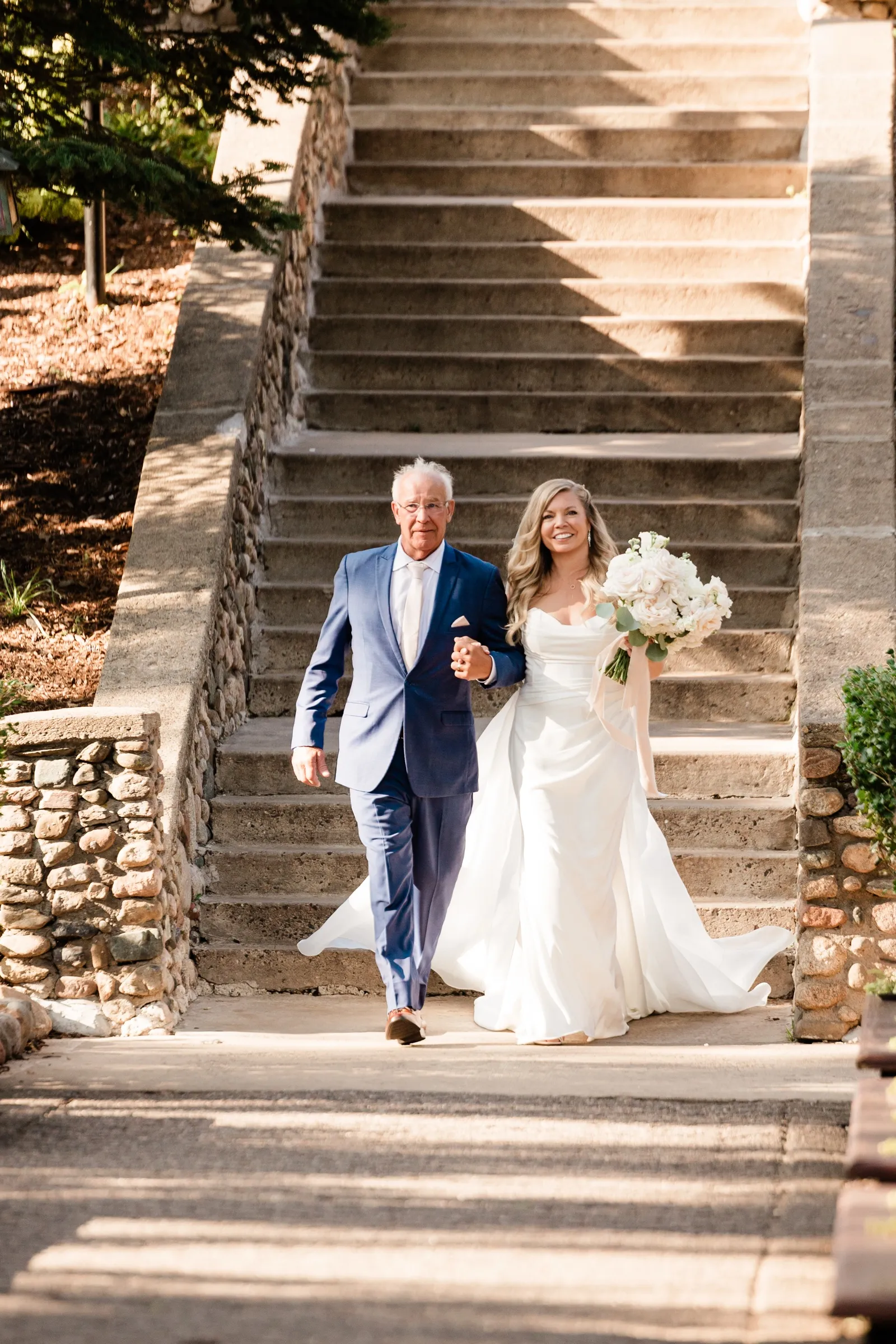 The bride and her father walk arm in arm down a wide stone staircase, her bouquet of white roses held at her side. — Tim Larsen Photography, Brainerd Lakes MN