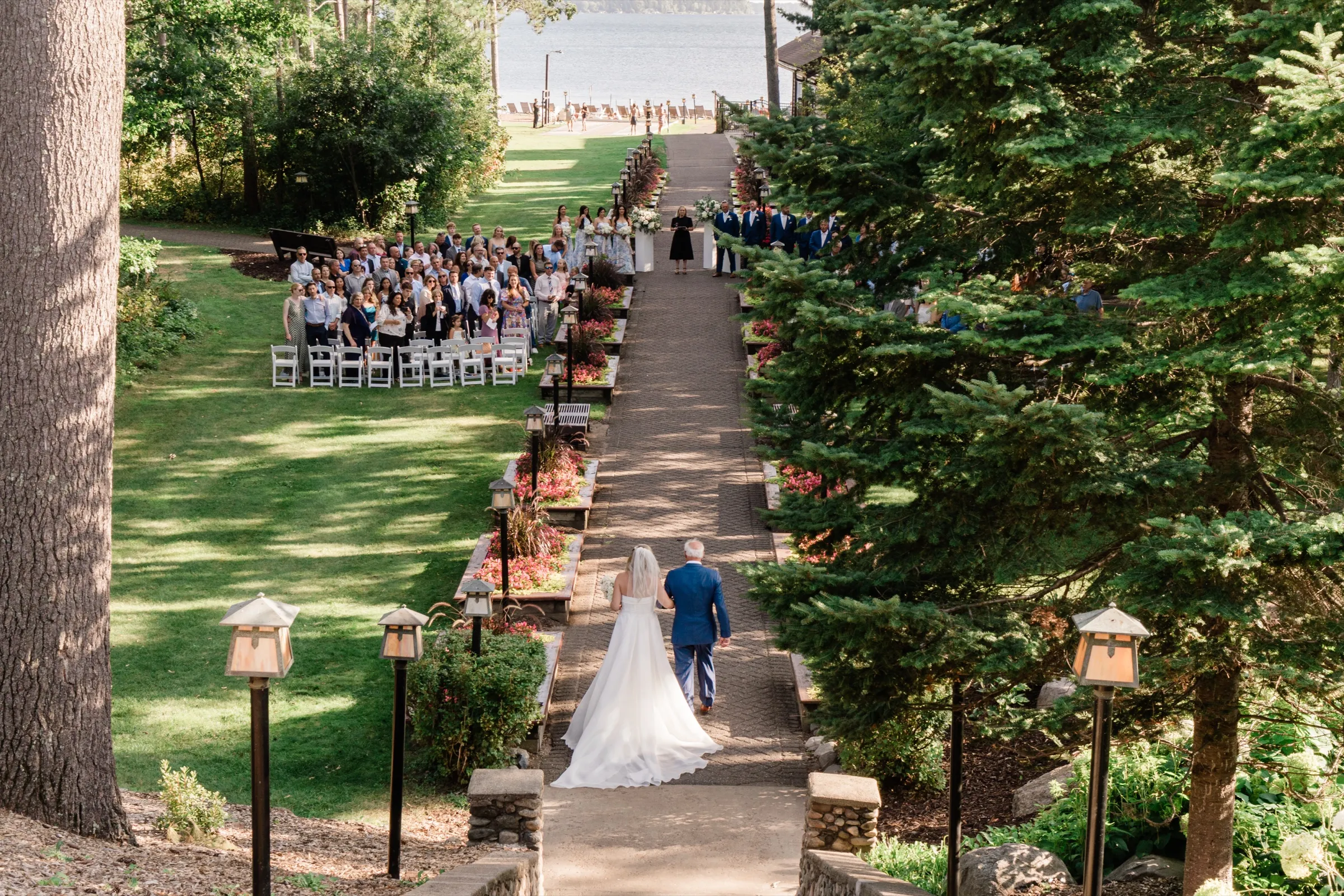 Seen from the top of a staircase, the bride and her father walk a lantern-lined path toward seated guests with Gull Lake beyond. — Tim Larsen Photography, Brainerd Lakes MN