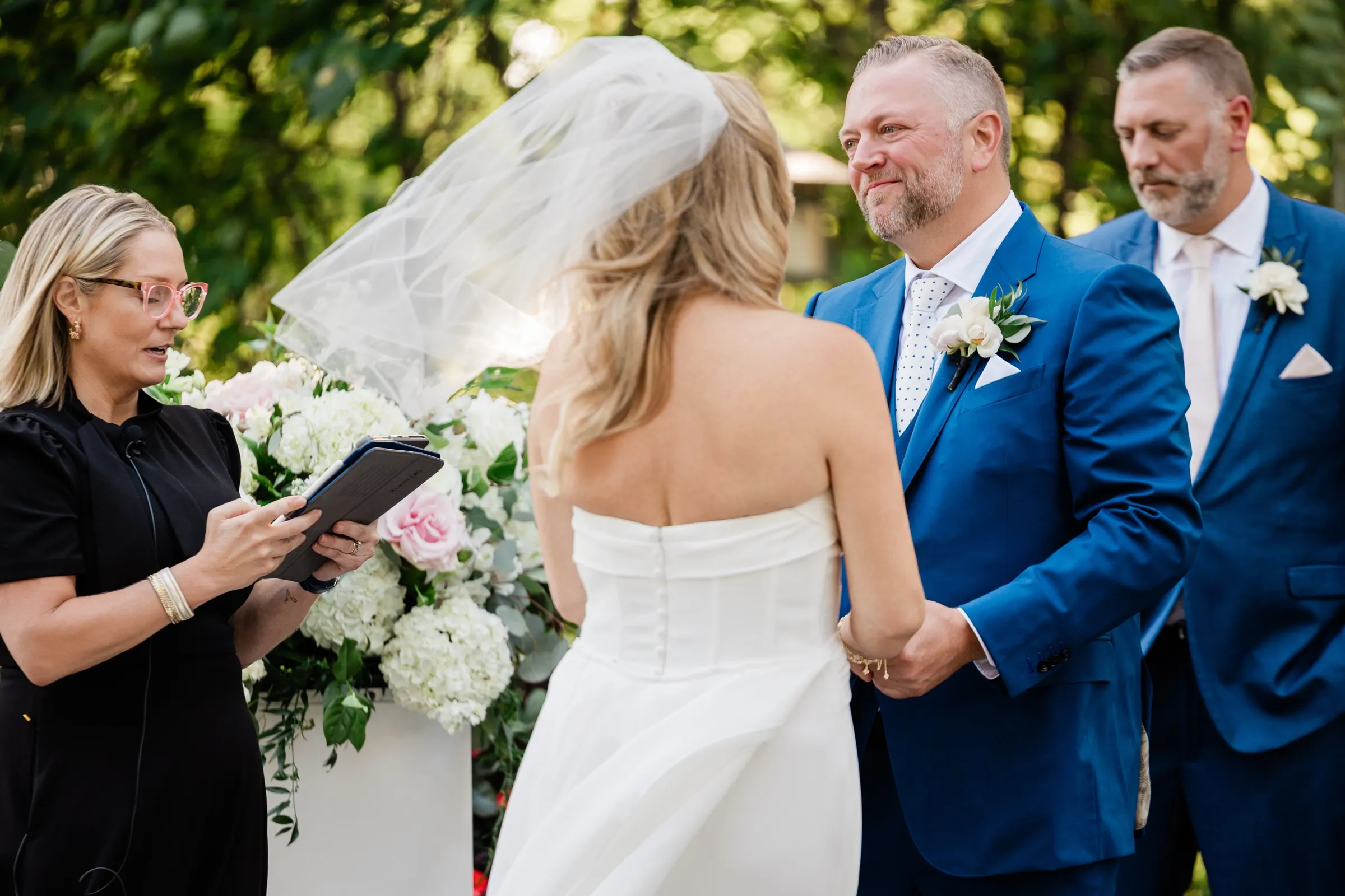 The groom holds the bride's hands and looks at her steadily while the officiant reads from a folder beside them. — Tim Larsen Photography, Brainerd Lakes MN