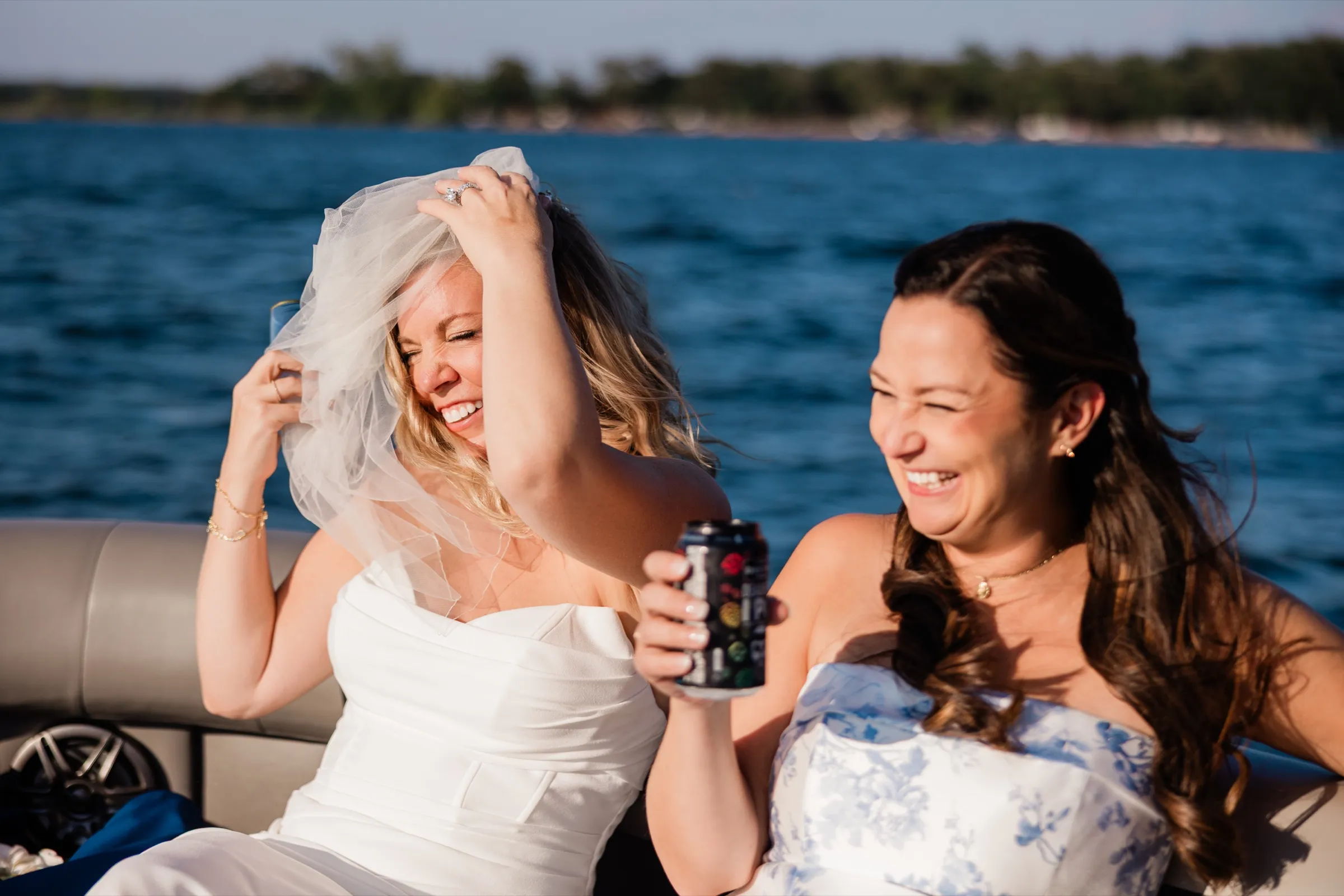 The bride grabs at her wind-blown veil and laughs with a friend on the back of a pontoon boat, deep blue water filling the background. — Tim Larsen Photography, Brainerd Lakes MN