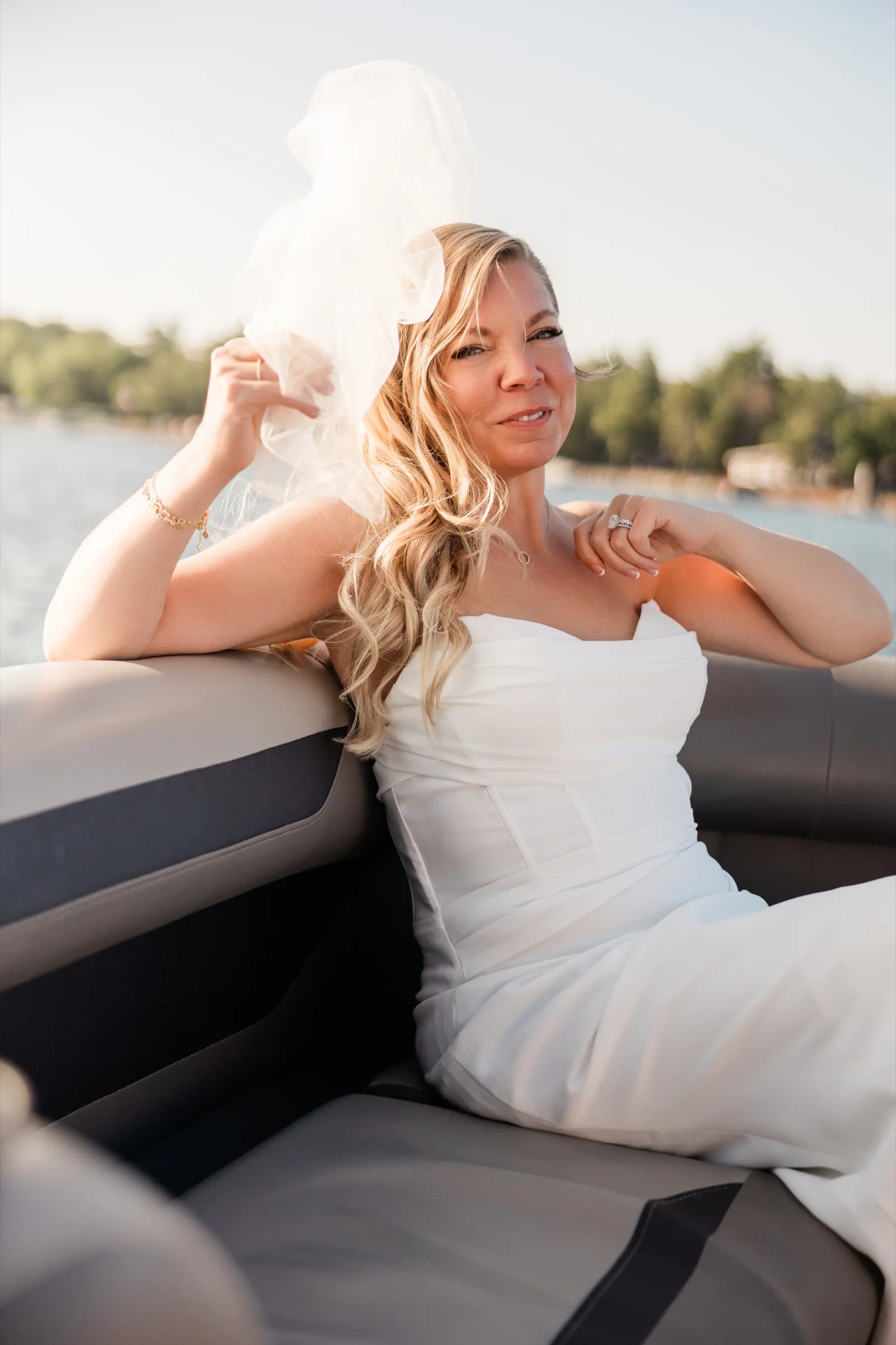 The bride leans back against a pontoon seat and holds her veil up in the wind, the far shoreline soft behind her. — Tim Larsen Photography, Brainerd Lakes MN