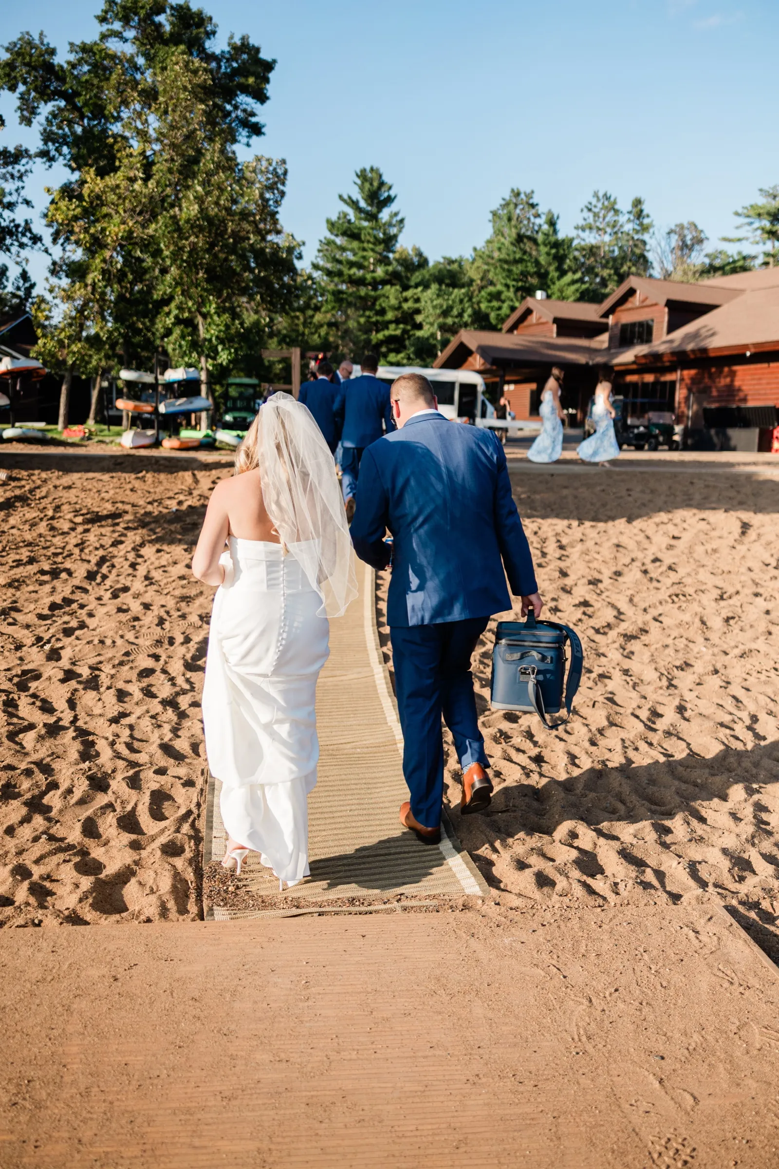 The bride and groom walk hand in hand up a mat across the sand toward the lodge buildings, seen from behind. — Tim Larsen Photography, Brainerd Lakes MN