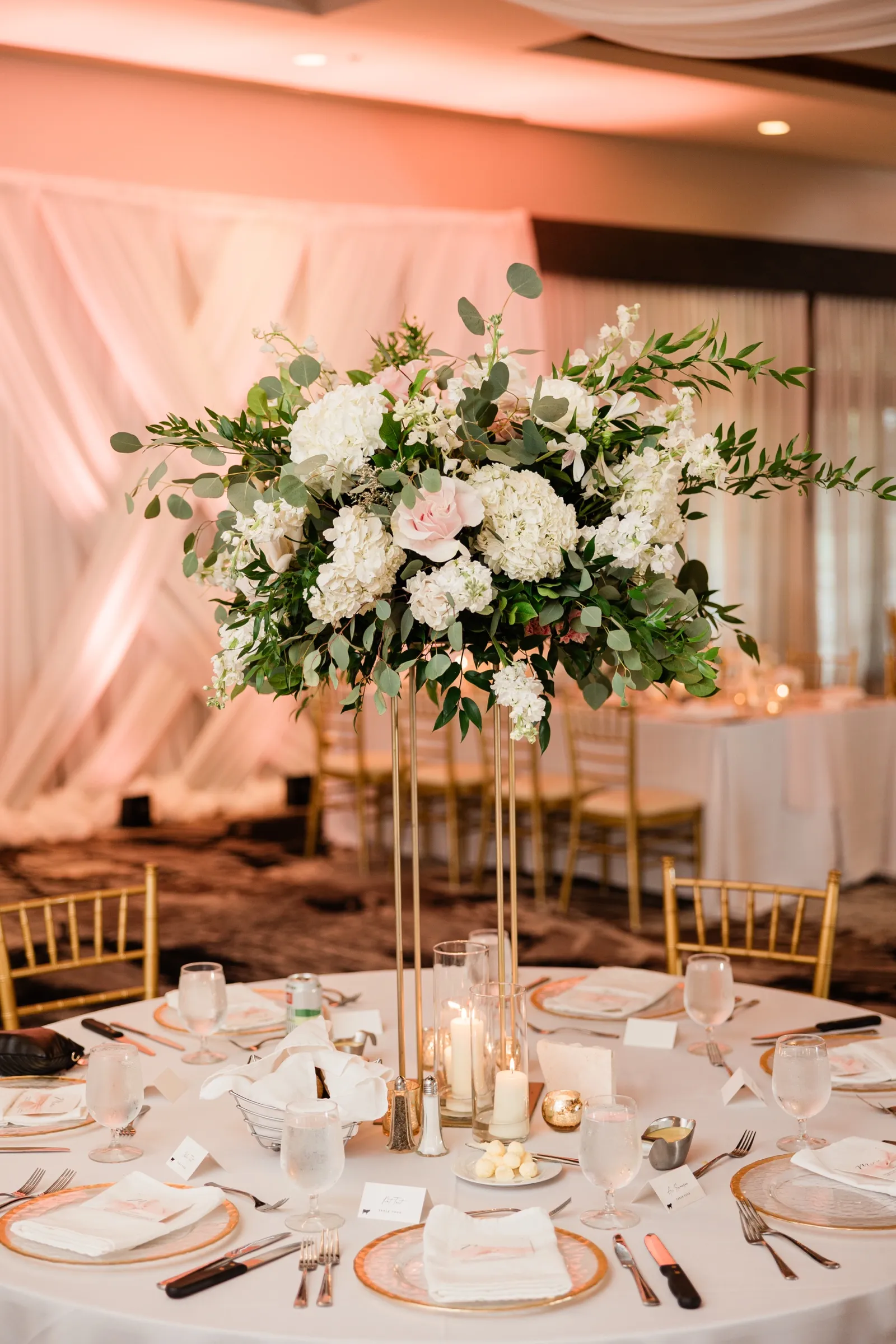 A tall white hydrangea and greenery centerpiece rises above a set round table in a blush-lit reception room. — Tim Larsen Photography, Brainerd Lakes MN