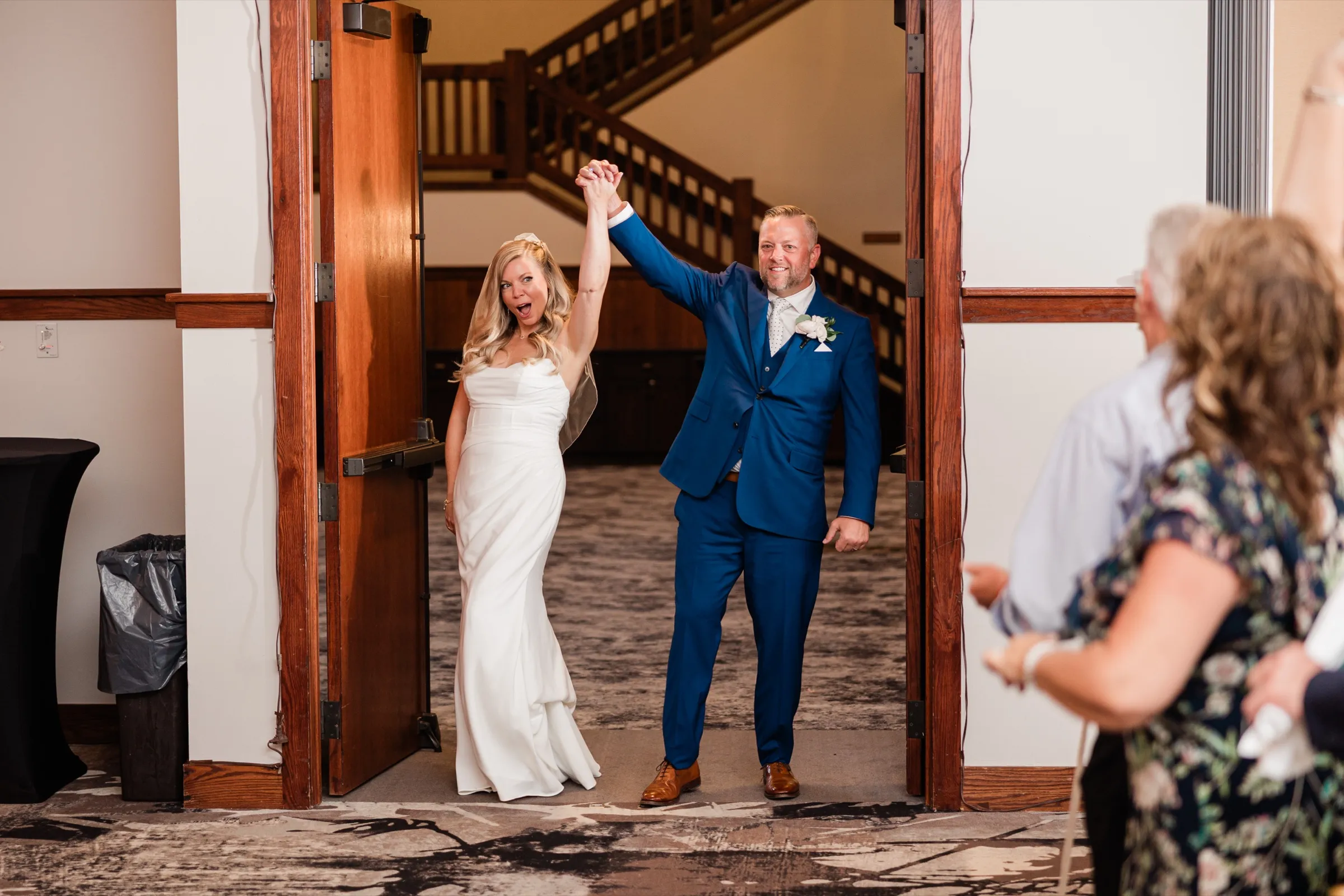 The bride and groom stand in an open doorway with their joined hands raised, a wooden staircase visible behind them. — Tim Larsen Photography, Brainerd Lakes MN