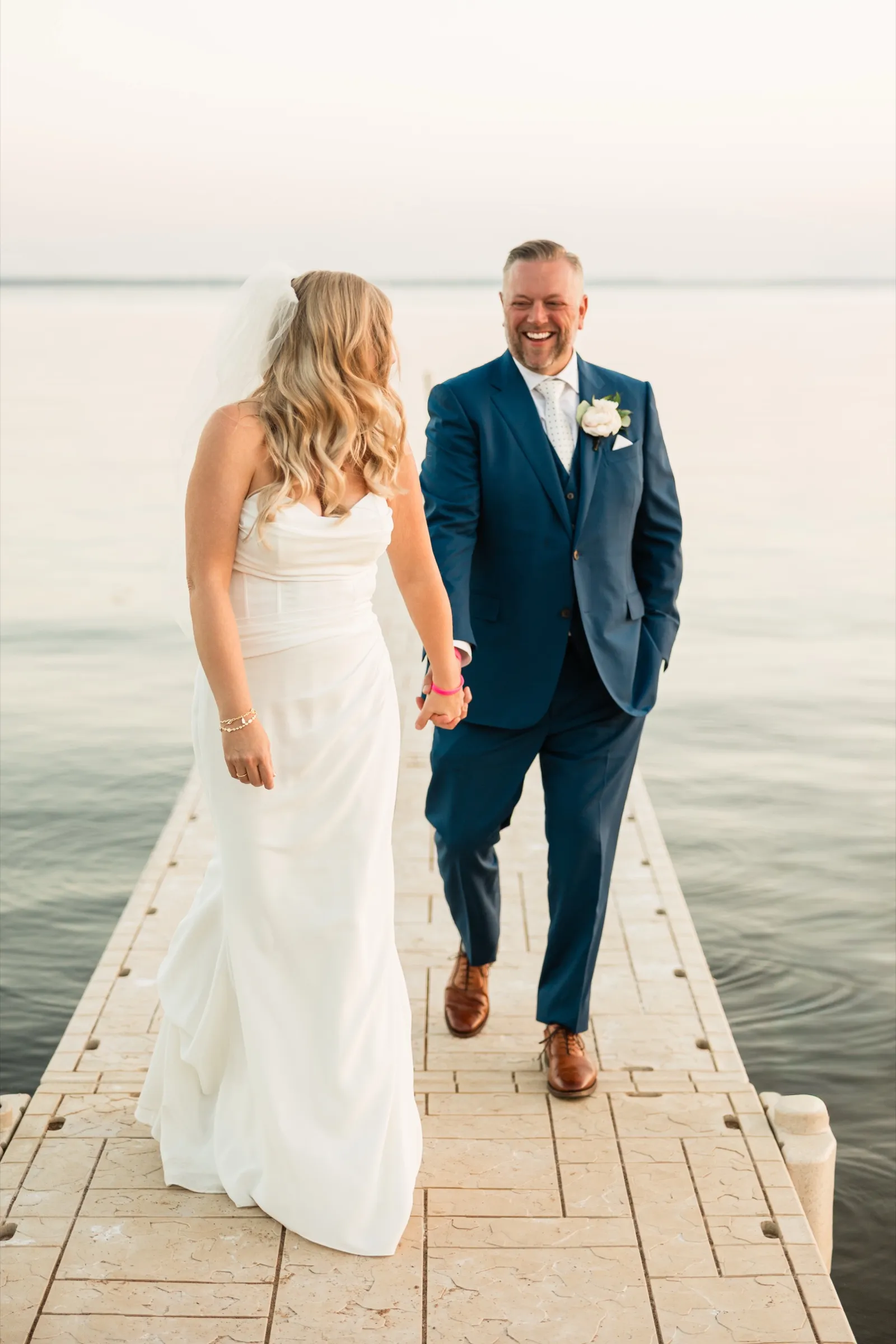 The groom walks toward the bride along a dock, laughing, as she holds his hand with her back to the camera. — Tim Larsen Photography, Brainerd Lakes MN