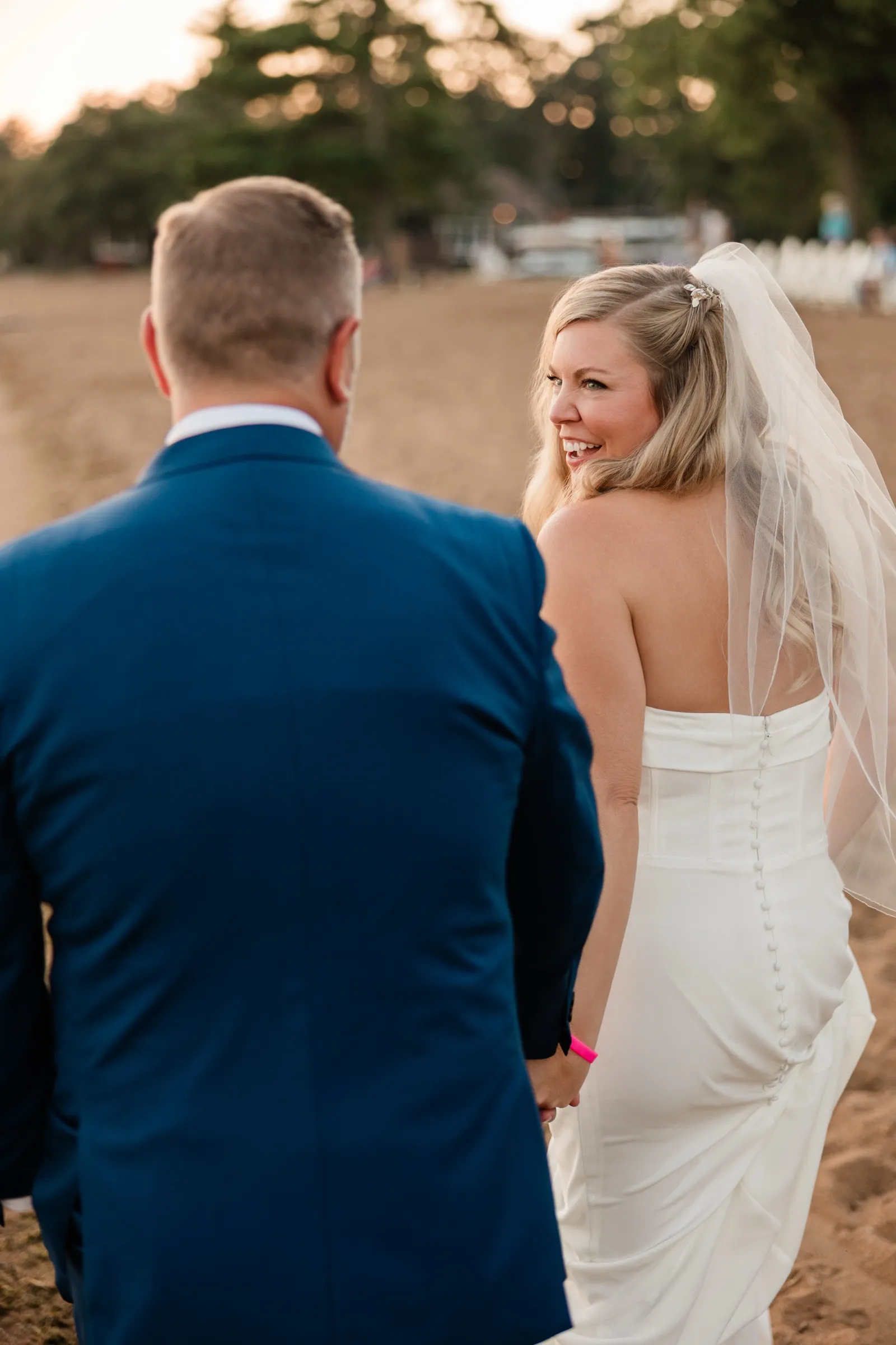 Walking away across the sand with the groom, the bride turns back over her shoulder laughing, her veil lit from behind. — Tim Larsen Photography, Brainerd Lakes MN