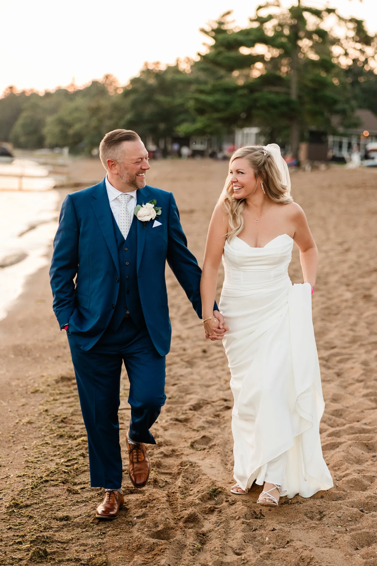 The bride and groom walk hand in hand across the sand, looking at each other and laughing, with pines behind them. — Tim Larsen Photography, Brainerd Lakes MN