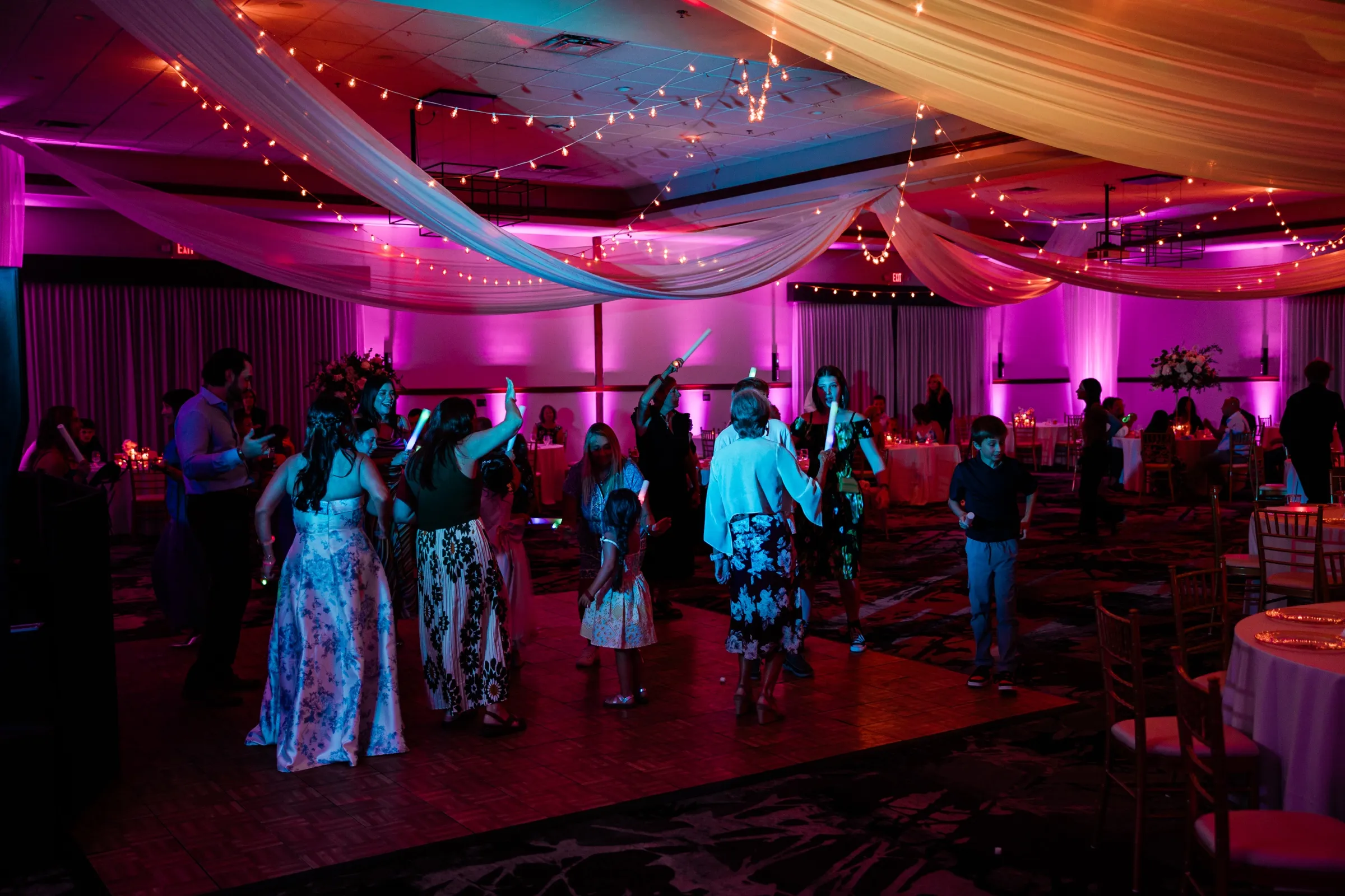 Guests dance with raised glow sticks across a wide ballroom lit magenta, with draped fabric and string lights overhead. — Tim Larsen Photography, Brainerd Lakes MN