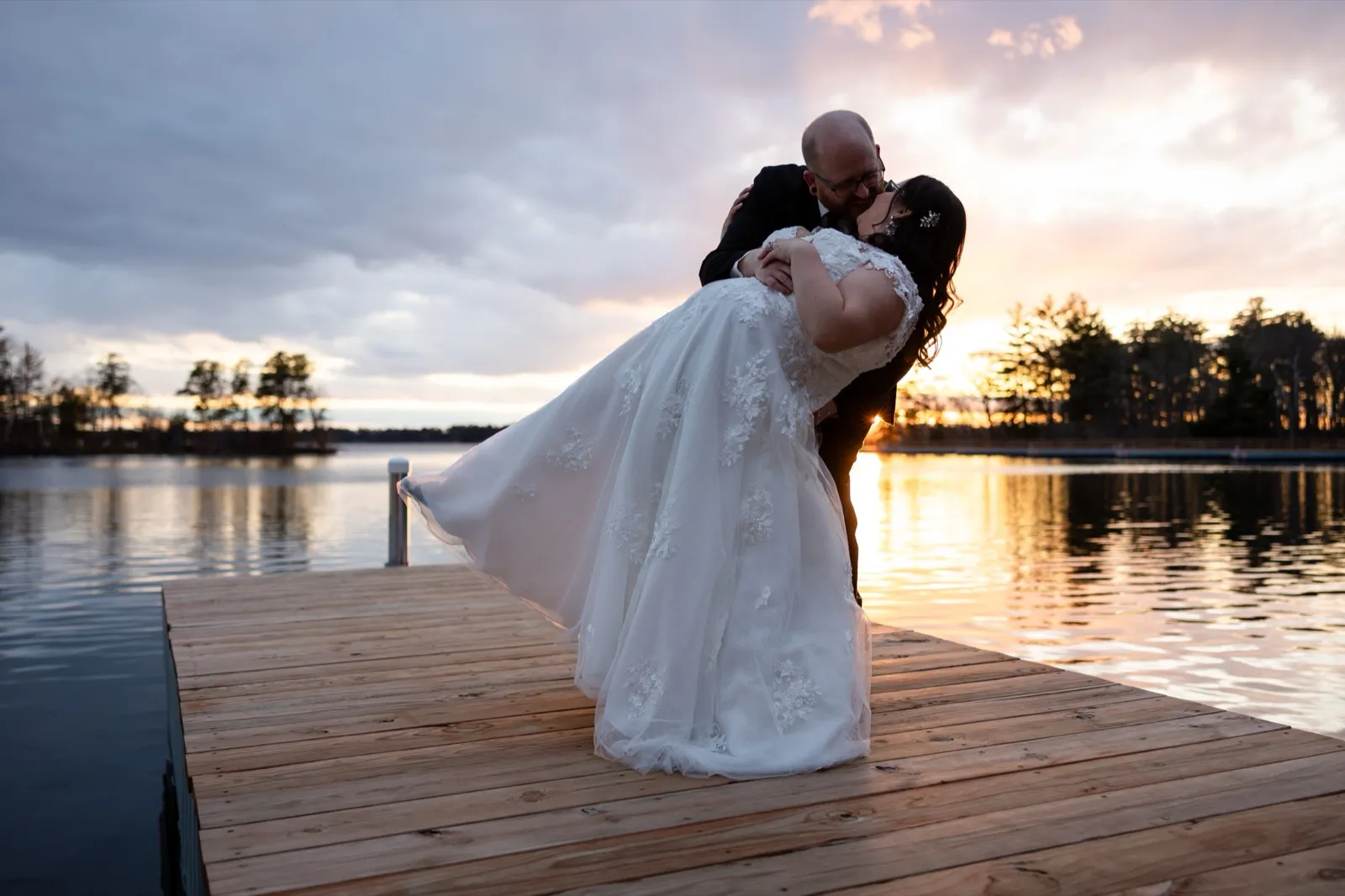 A groom dips the bride for a kiss at the end of a dock as the sun sets over the lake behind them, the last warm light on the water — Tim Larsen Photography, Brainerd Lakes MN