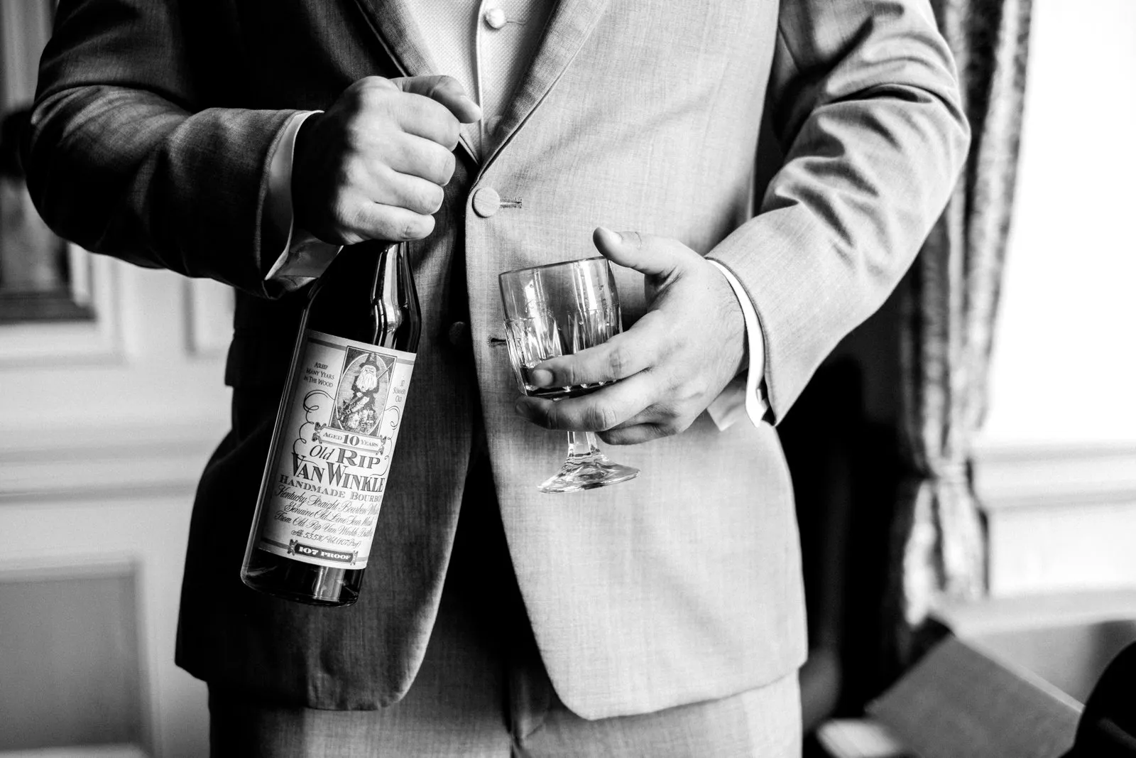 Tight crop on the groom’s suit and vest with a bourbon bottle and glass framing his buttoning hands — Greysolon Plaza getting-ready detail — Tim Larsen Photography, Duluth MN