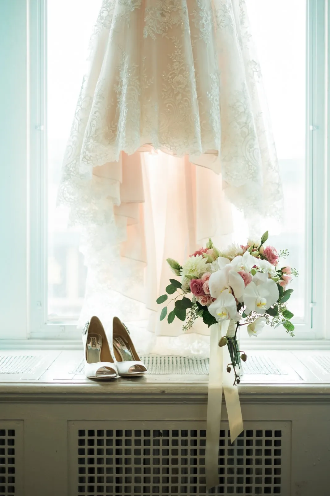 Bridal gown backlit in the tall arched window of the Greysolon Plaza bridal suite, shoes and cascading orchid bouquet anchored on the radiator sill below — Tim Larsen Photography, Duluth MN