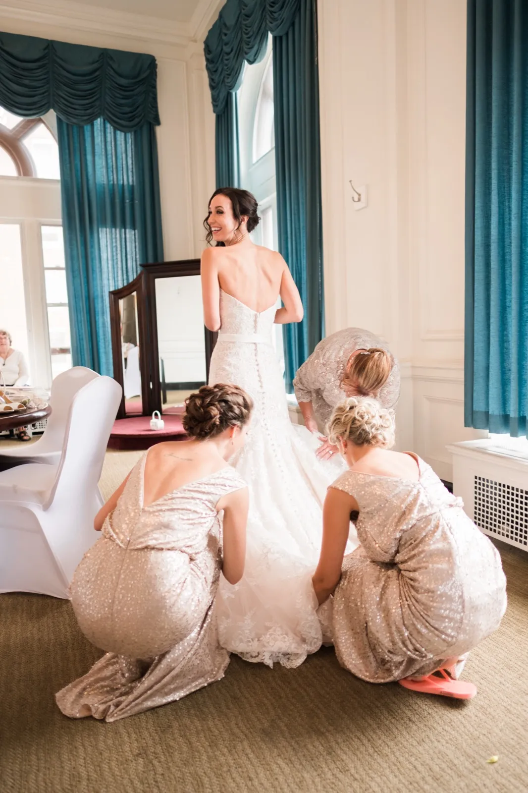 Bride glances back smiling while three crouched bridesmaids fan her gown’s train in the Greysolon Plaza bridal suite — Tim Larsen Photography, Duluth MN