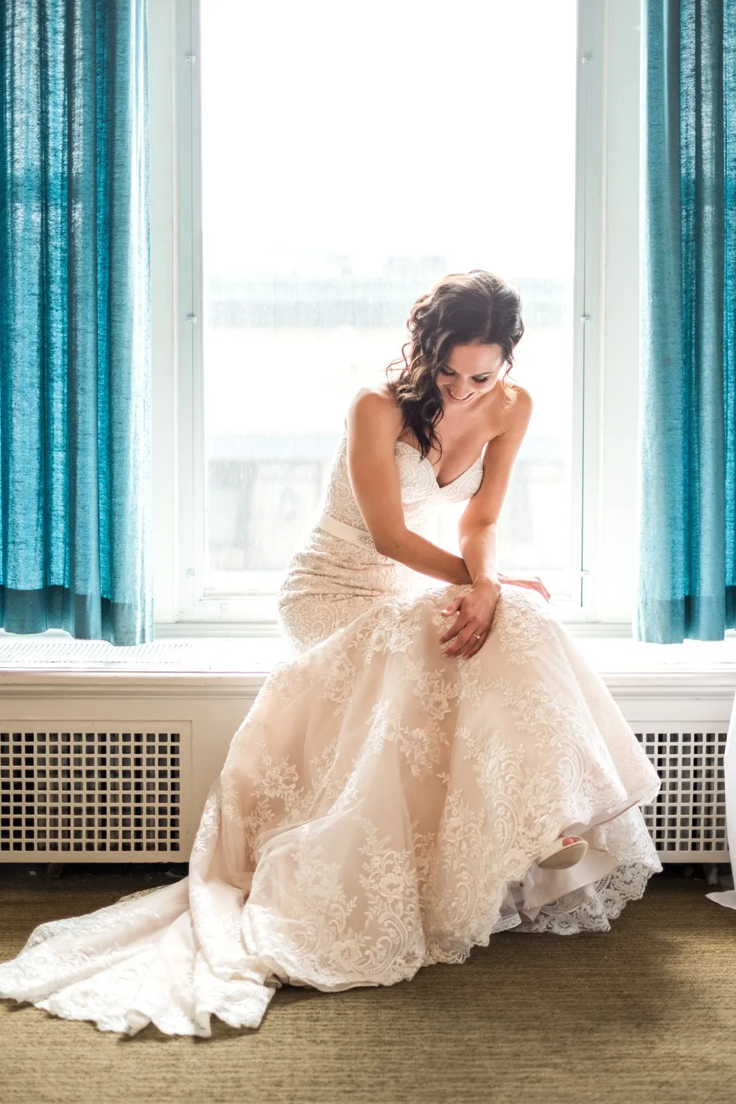 Centered bridal portrait framed in the tall arched window of the Greysolon bridal suite, lace train fanned across the floor and teal velvet curtains framing the room — Tim Larsen Photography, Duluth MN