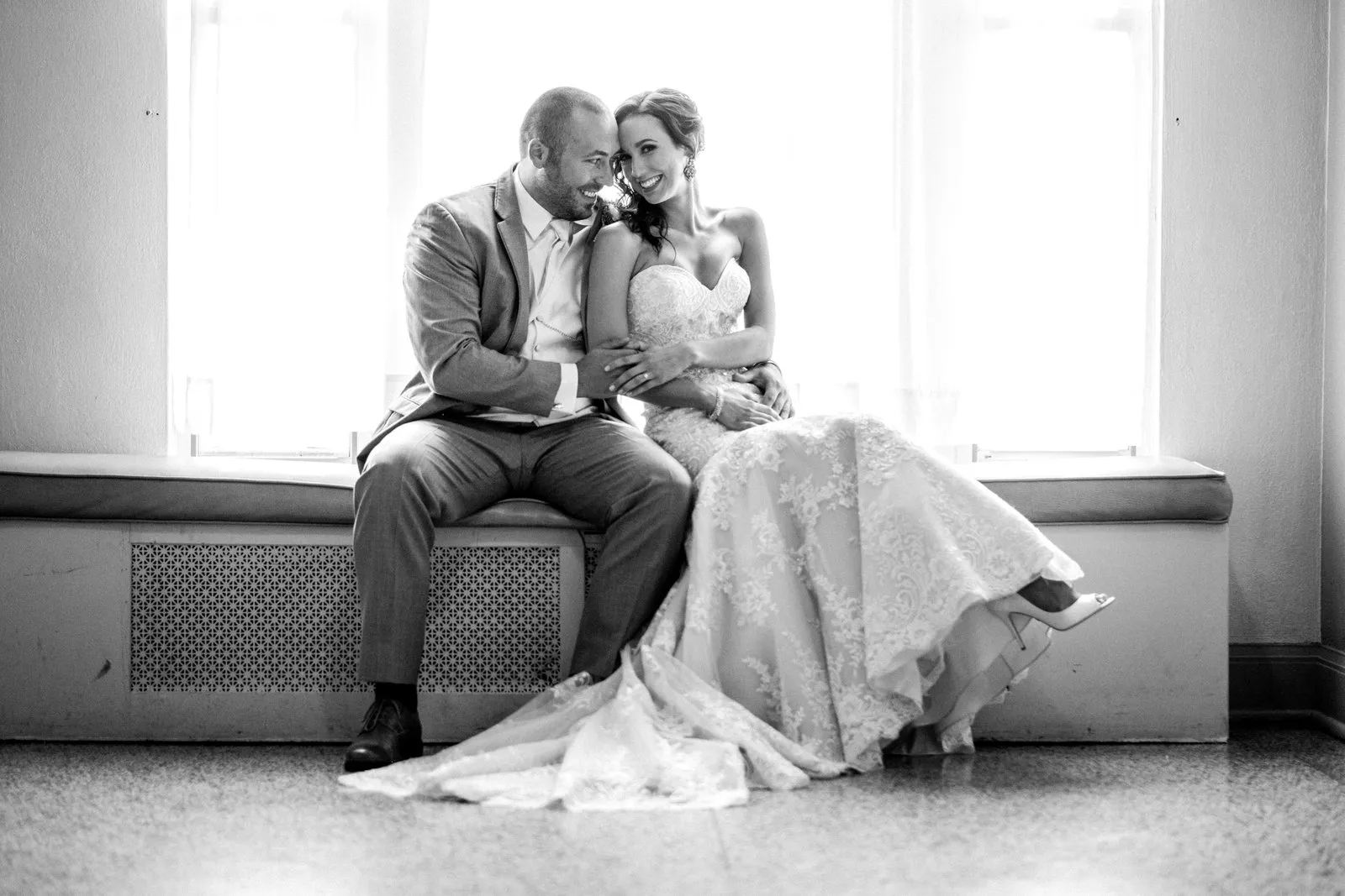 Symmetrical window-backlit couples portrait — bride and groom centered on a radiator bench in the Greysolon Plaza, lace gown spilling diagonally across the floor — Tim Larsen Photography, Duluth MN