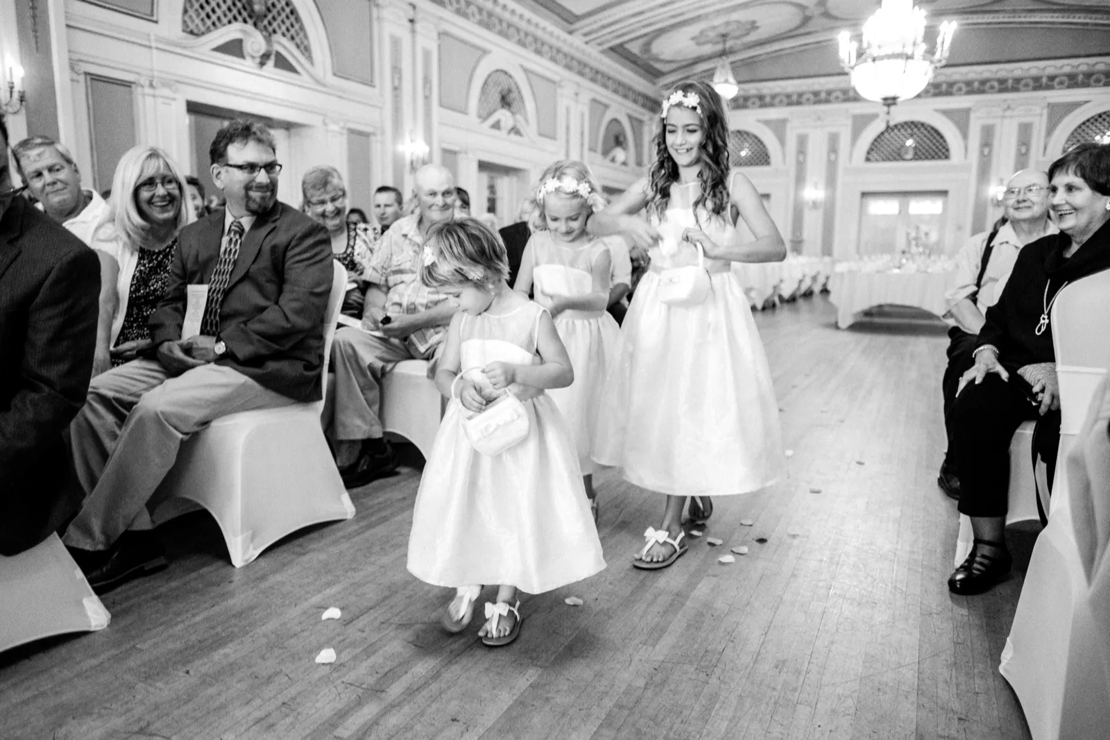Three flower girls walk mid-aisle as grinning guests flank them, the ornate ballroom ceiling arching above — Greysolon Ballroom ceremony processional — Tim Larsen Photography, Duluth MN