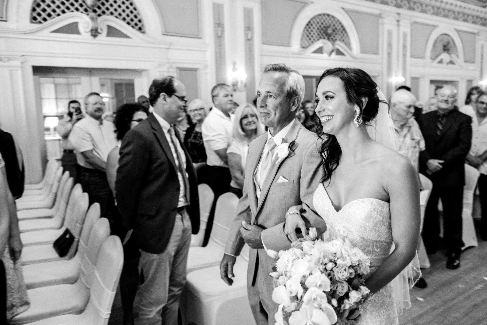 Bride and her father walk down the Greysolon Ballroom aisle as seated guests turn to watch, ornate ballroom architecture framing the processional — Tim Larsen Photography, Duluth MN