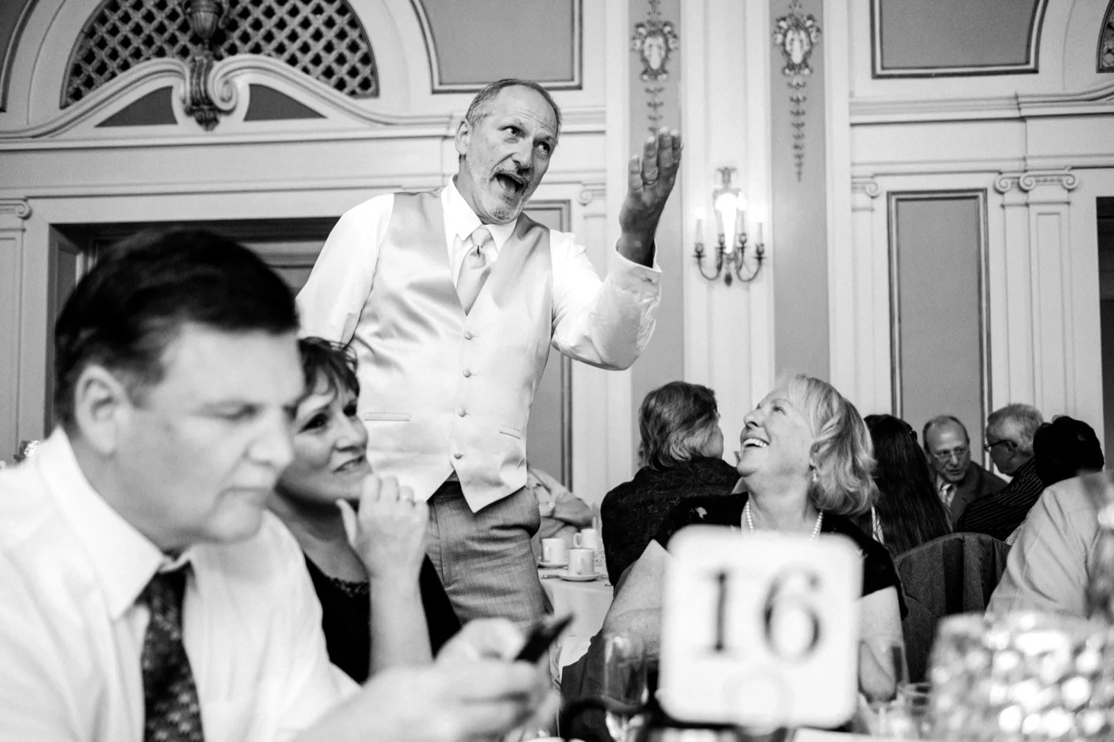 Reception candid — standing guest reaches upward mid-story while a seated woman laughs, foreground figure soft, ornate Greysolon Ballroom architecture framing the moment — Tim Larsen Photography, Duluth MN