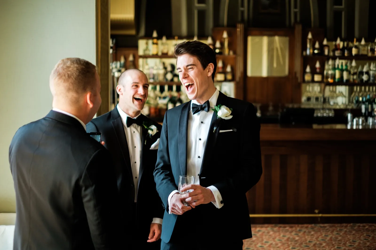 The groom laughs open-mouthed beside two groomsmen at an ornate wood bar during cocktail hour inside the Greysolon Plaza — Tim Larsen Photography, Duluth MN