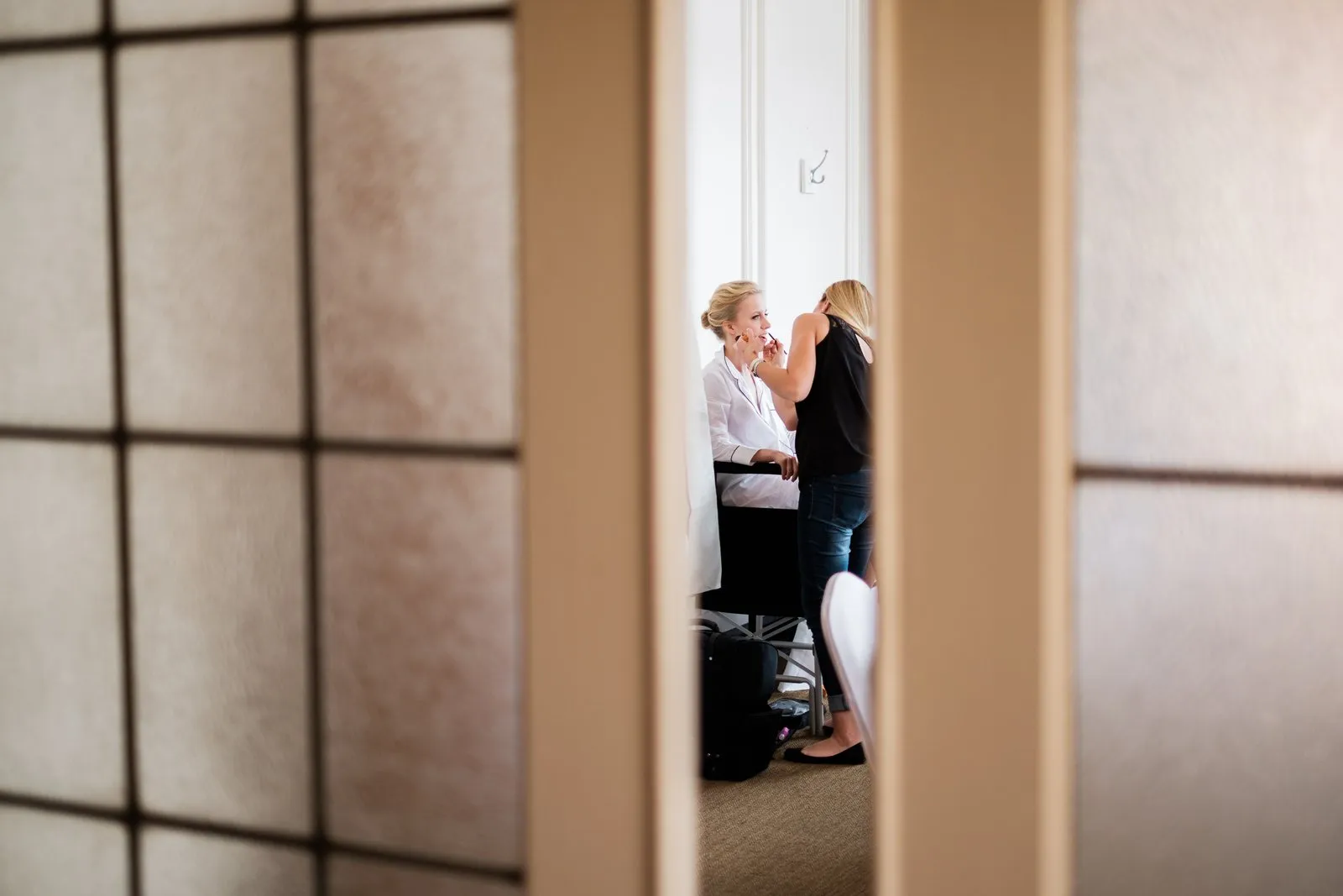 The bride sits in window light having her makeup applied, viewed through the narrow opening of a shoji-style sliding door — Greysolon Plaza bridal suite — Tim Larsen Photography, Duluth MN