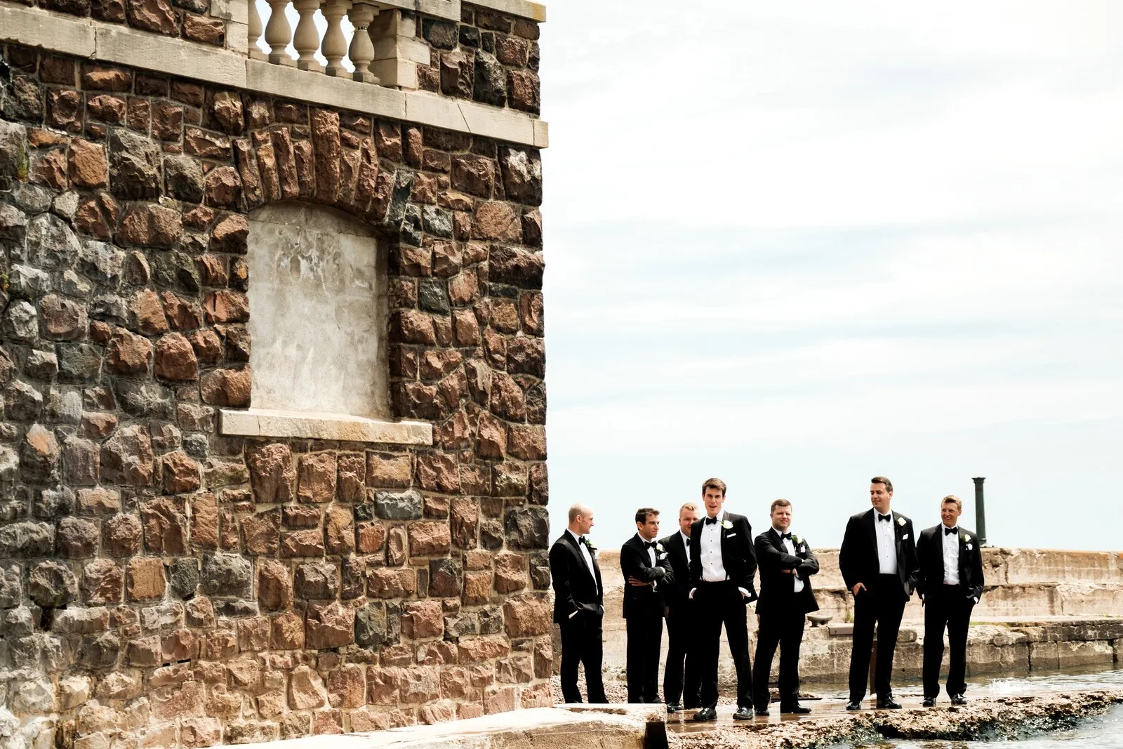 Groomsmen in black tuxedos gather against the textured stone wall and arched balustrade of Greysolon Plaza, Lake Superior visible at right — Tim Larsen Photography, Duluth MN