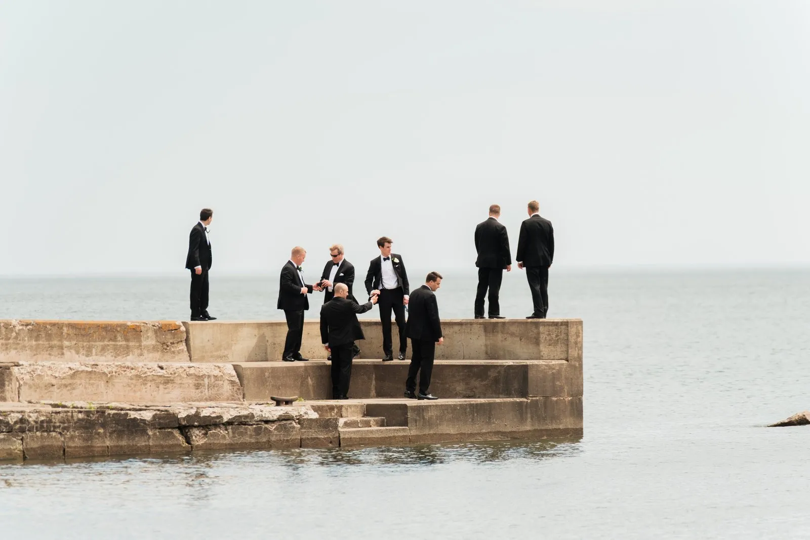 Eight groomsmen in black tuxedos stand scattered across a concrete pier extending into Lake Superior under an overcast sky — Tim Larsen Photography, Duluth MN