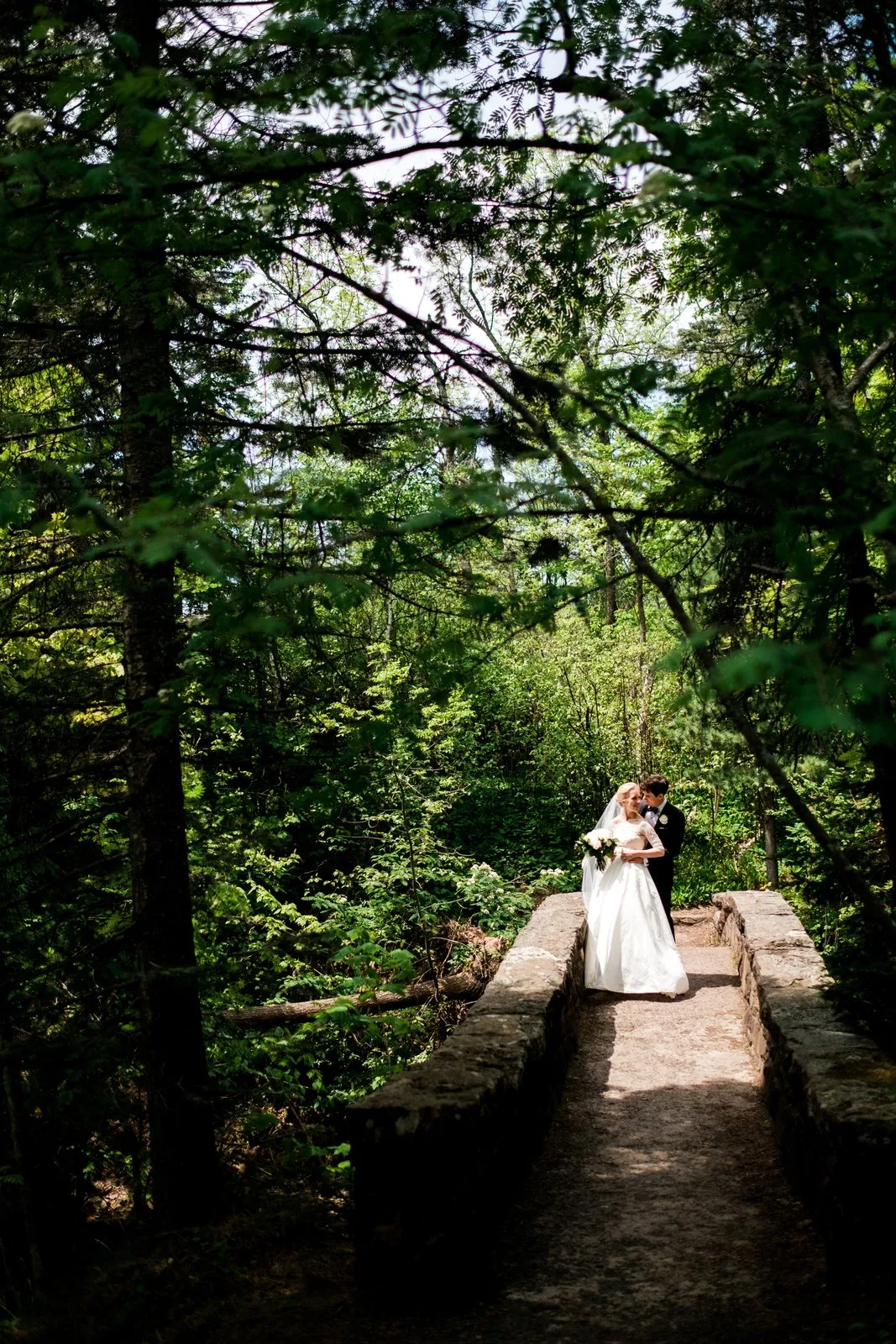 The bride and groom embrace on a sunlit stone footbridge, framed by an arching tunnel of dense green forest foliage — Tim Larsen Photography, Duluth MN
