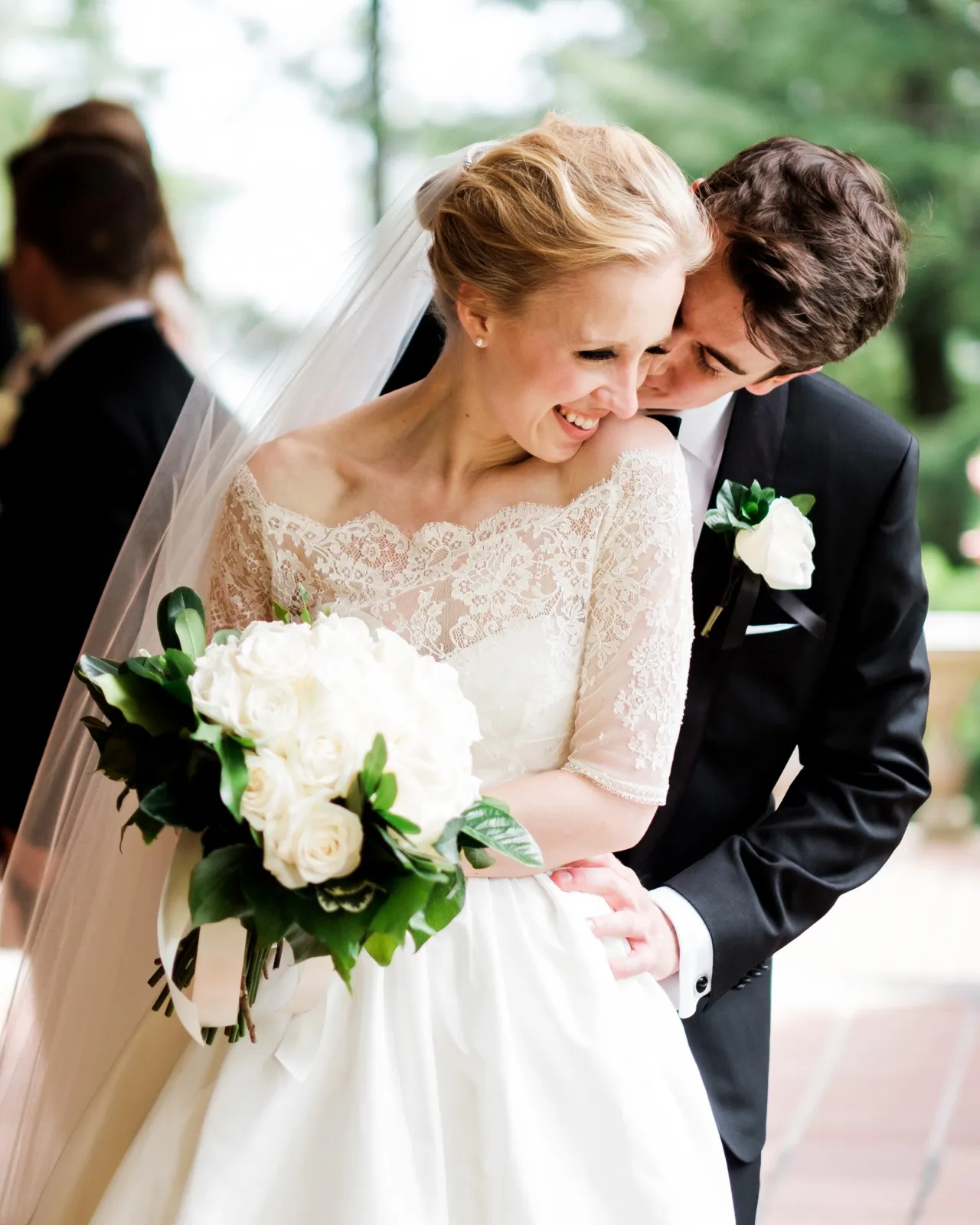 The groom nuzzles into the laughing bride's cheek as she holds her white rose bouquet, her lace sleeves and veil catching soft outdoor light — Tim Larsen Photography