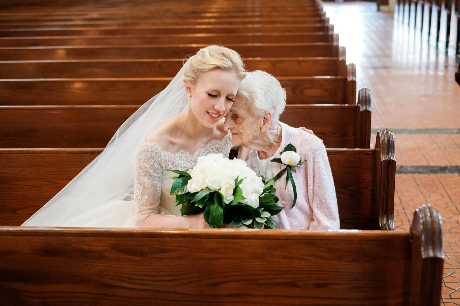 A bride leans her forehead against her elderly grandmother's as they sit together in a church pew, the grandmother in soft pink, the bride's bouquet of white roses resting between them — Tim Larsen Photography, Brainerd Lakes MN