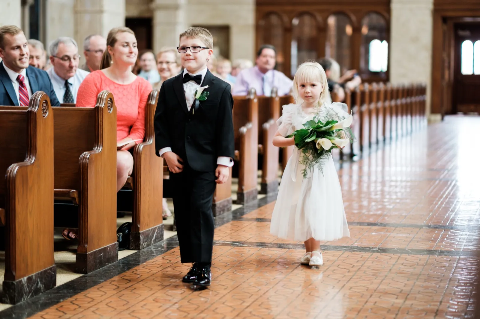 A young ring bearer in a black tuxedo and a flower girl in a white dress holding a bouquet walk down the cathedral aisle past seated guests in wooden pews — Tim Larsen Photography, Duluth MN