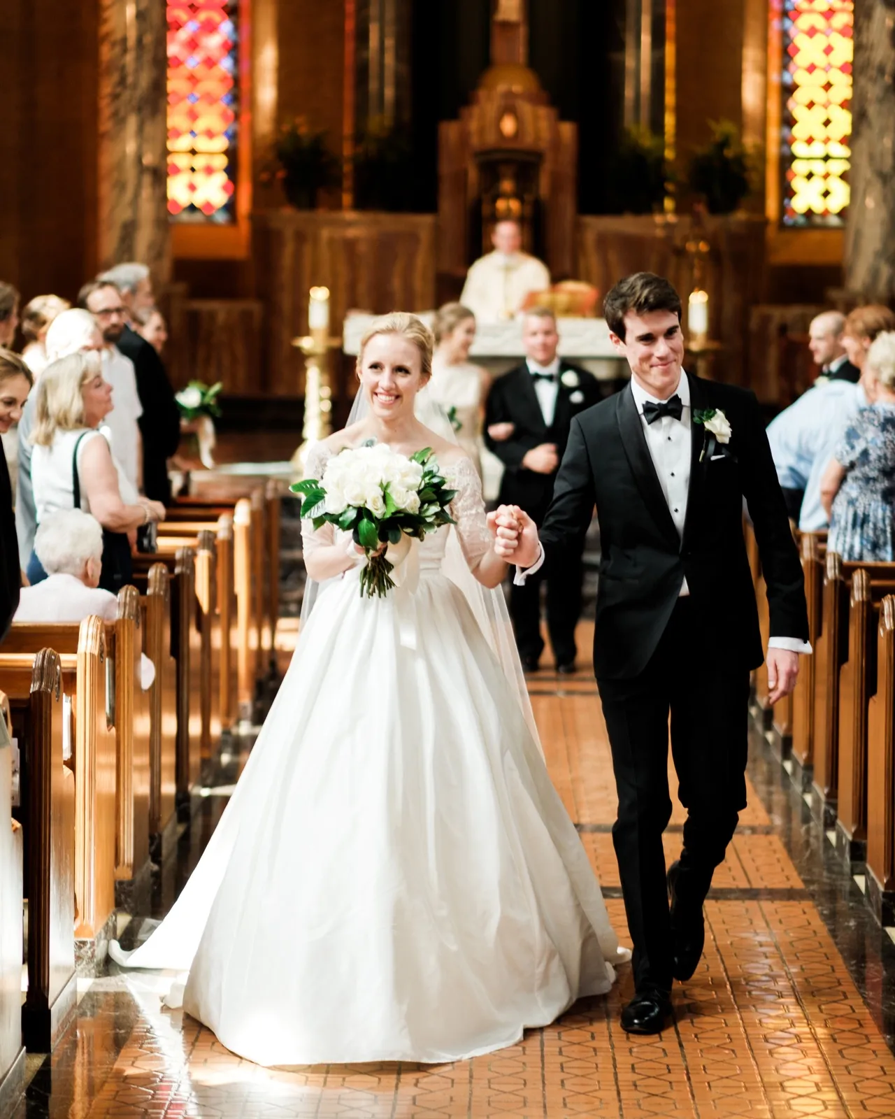 The bride and groom walk back up the cathedral aisle hand in hand just after their ceremony, both smiling, as guests rise in the pews behind them — Tim Larsen Photography