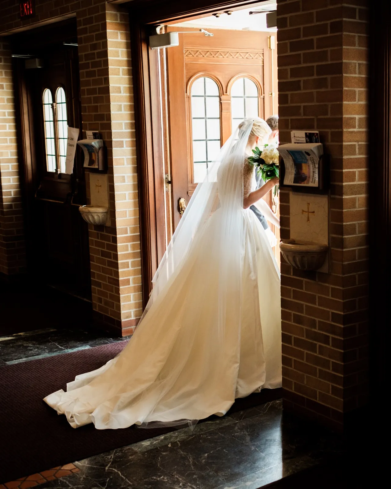 The bride seen from behind, her veil and gown train trailing across the floor as she walks toward the carved wooden doors of the Cathedral of Our Lady of the Rosary — Tim Larsen Photography