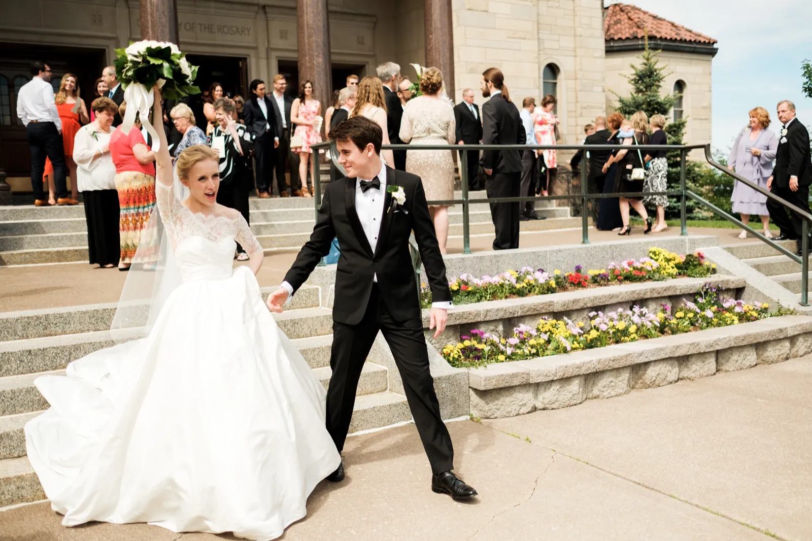 The bride raises her bouquet overhead in triumph as the groom leads her down the stone Cathedral steps past rows of cheering guests and beds of bright flowers — Tim Larsen Photography, Duluth MN