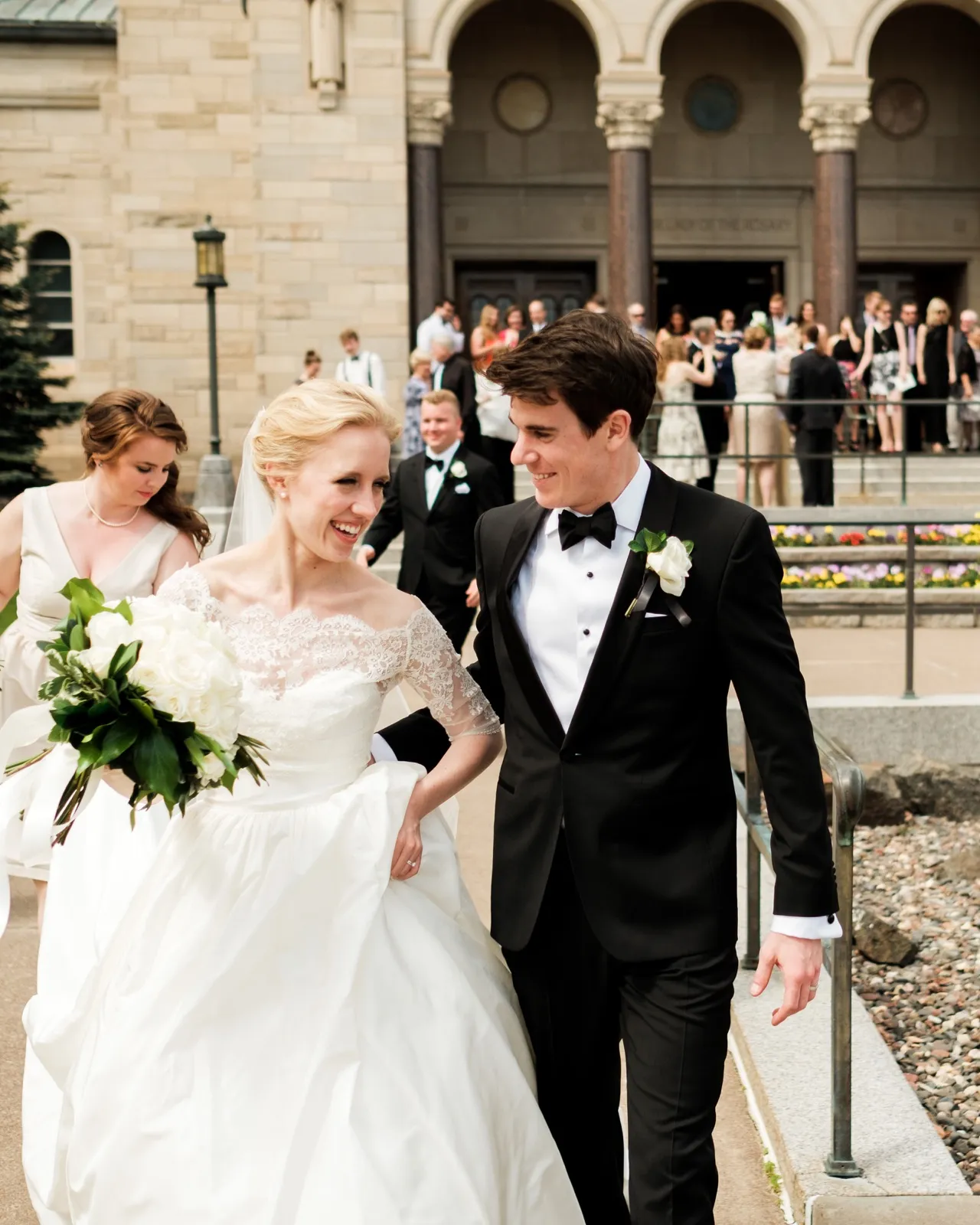 The bride laughs beside the groom on the Cathedral steps just after the ceremony, a bridesmaid and cheering guests behind them — Tim Larsen Photography