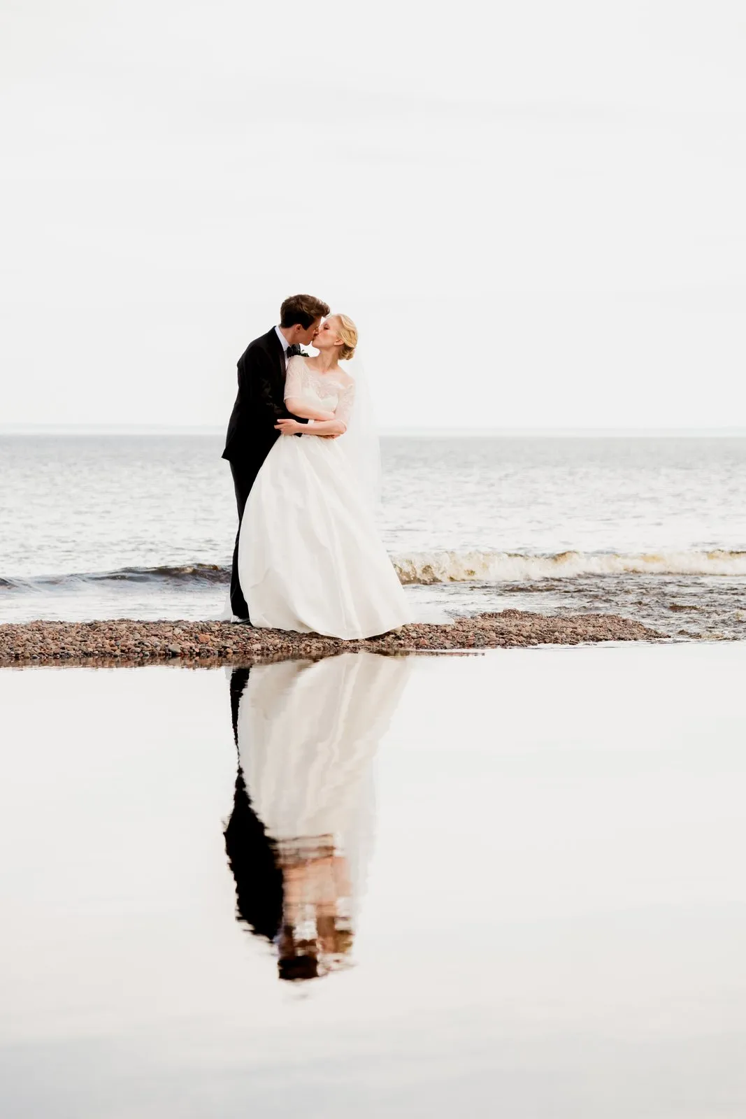 The bride and groom kiss on the rocky Lake Superior shore, their full figures perfectly mirrored in a still tidal pool in the foreground — Tim Larsen Photography, Duluth MN