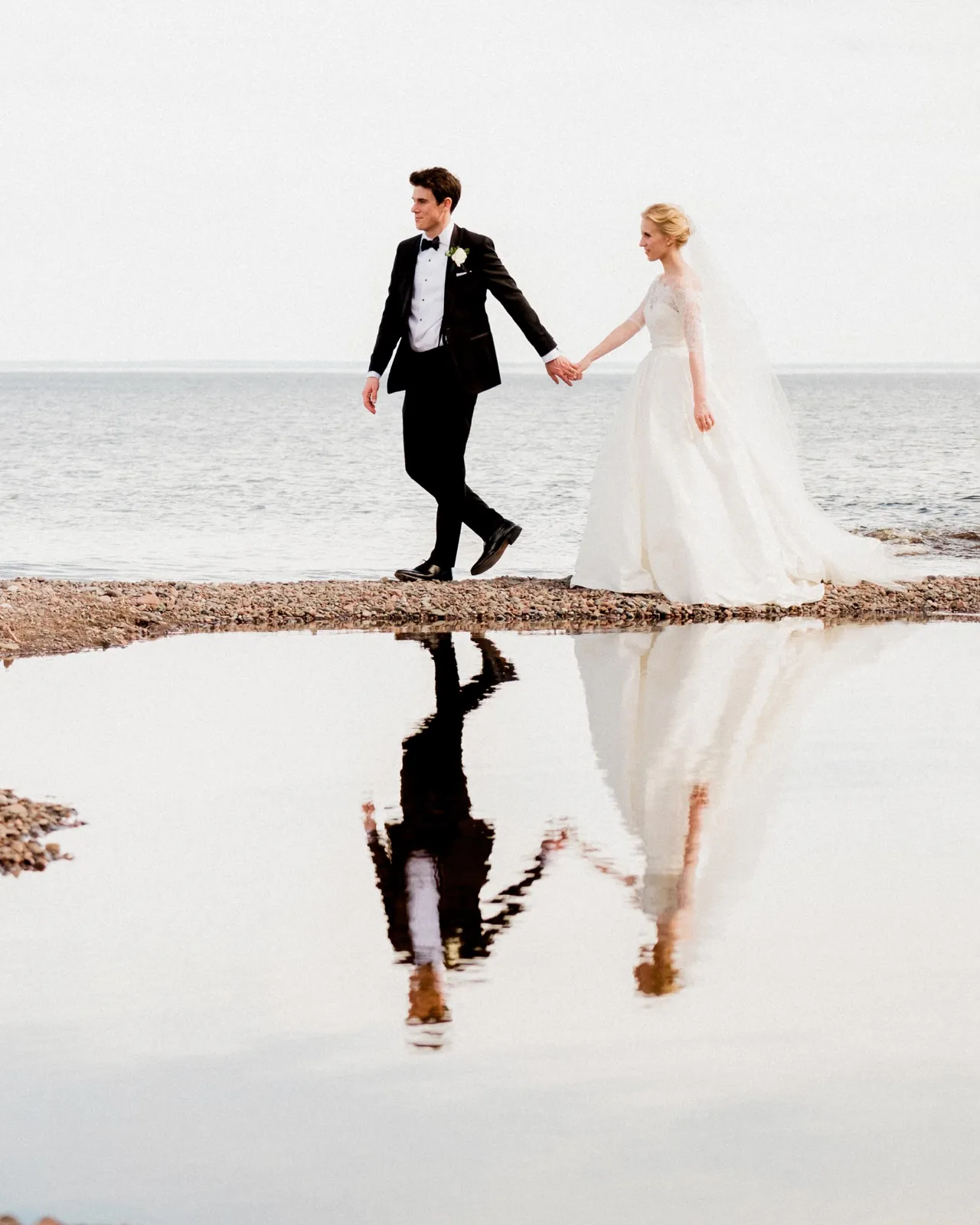 The bride and groom walk hand in hand along the rocky Lake Superior shoreline in Duluth, their figures mirrored in a still tidal pool below — Tim Larsen Photography