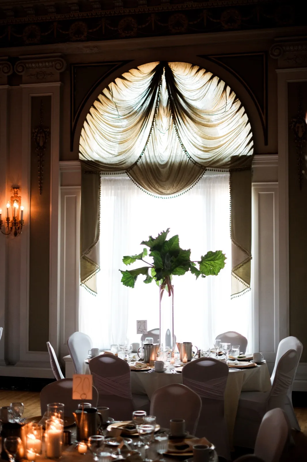 A tall rhubarb-leaf centerpiece rises in front of a grand draped arched window in the Greysolon Ballroom above set reception tables and chair-covered seats — Tim Larsen Photography, Duluth MN