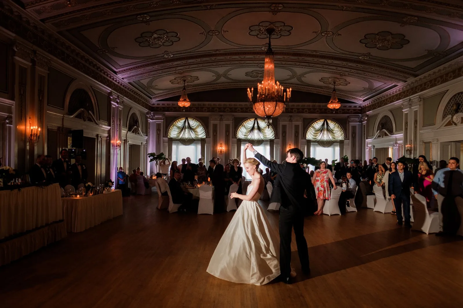The groom twirls the bride beneath the grand chandeliers of the Greysolon Ballroom, her skirt fanning wide as seated guests watch from the perimeter — Duluth wedding photography — Tim Larsen Photography, Duluth MN