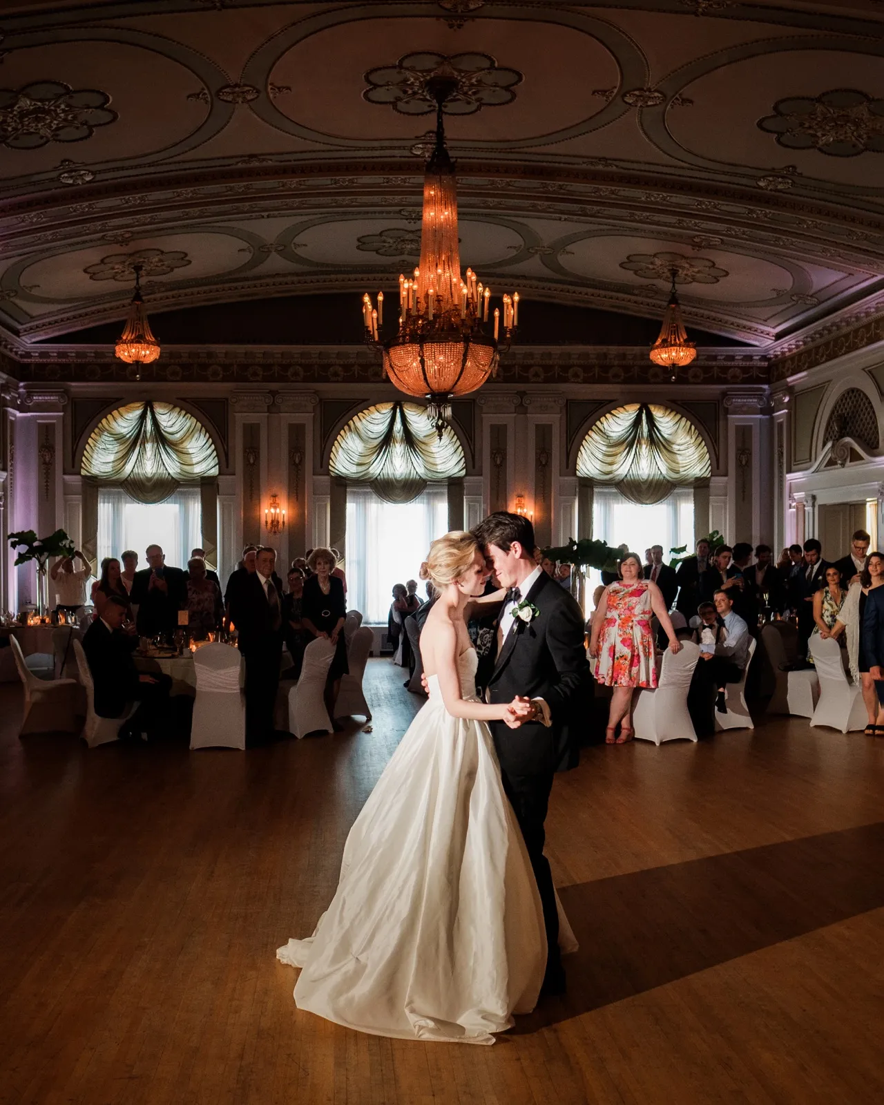 The bride and groom share their first dance beneath the grand chandelier and frescoed ceiling of the Greysolon Ballroom as guests watch from the edges of the floor — Tim Larsen Photography