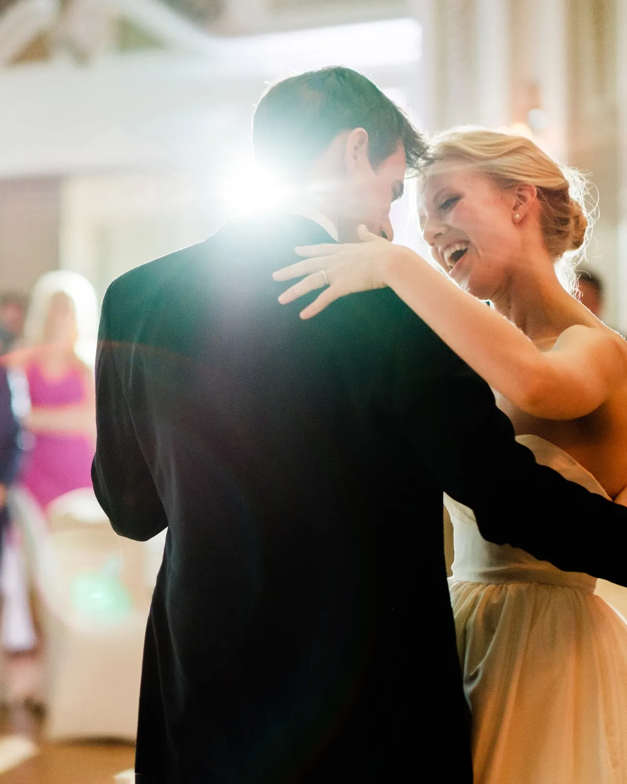 The bride laughs with her arms around the groom during their first dance, backlit by a warm flare on the Greysolon Ballroom floor — Tim Larsen Photography