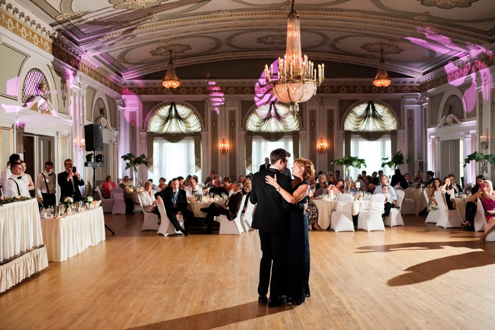 The groom dances with his mother on the empty ballroom floor beneath an ornate chandelier as seated guests watch from tables ringing the historic Greysolon Ballroom — Tim Larsen Photography, Duluth MN