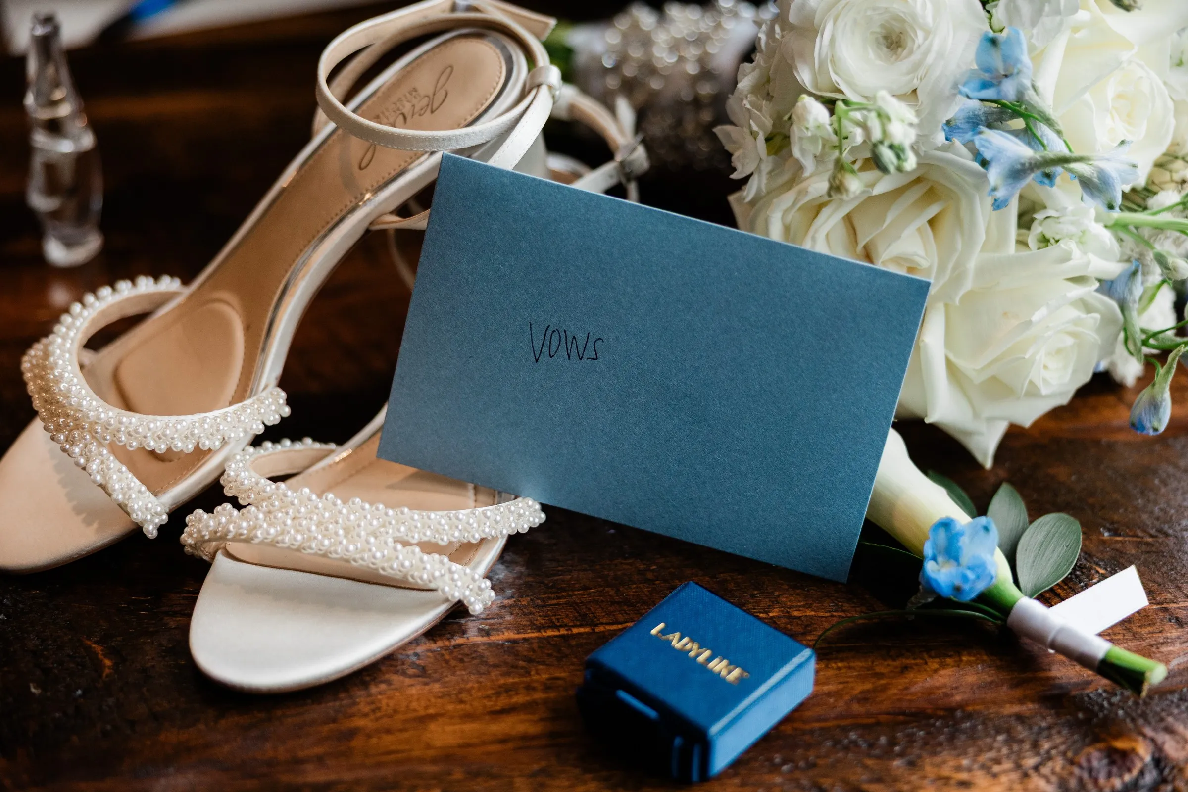 Pearl-trimmed bridal shoes, a blue vow book, rings and a white and blue bouquet arranged on a wooden table — Tim Larsen Photography, Brainerd Lakes MN