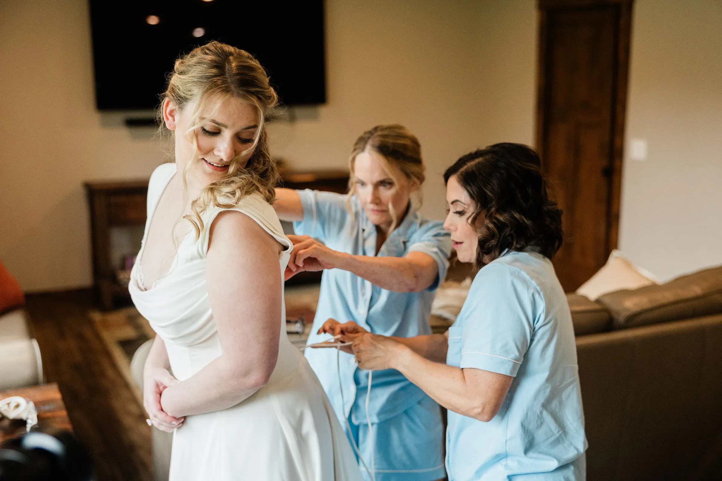 Two women help fasten Lauren’s wedding gown as she looks over her shoulder — Tim Larsen Photography, Brainerd Lakes MN