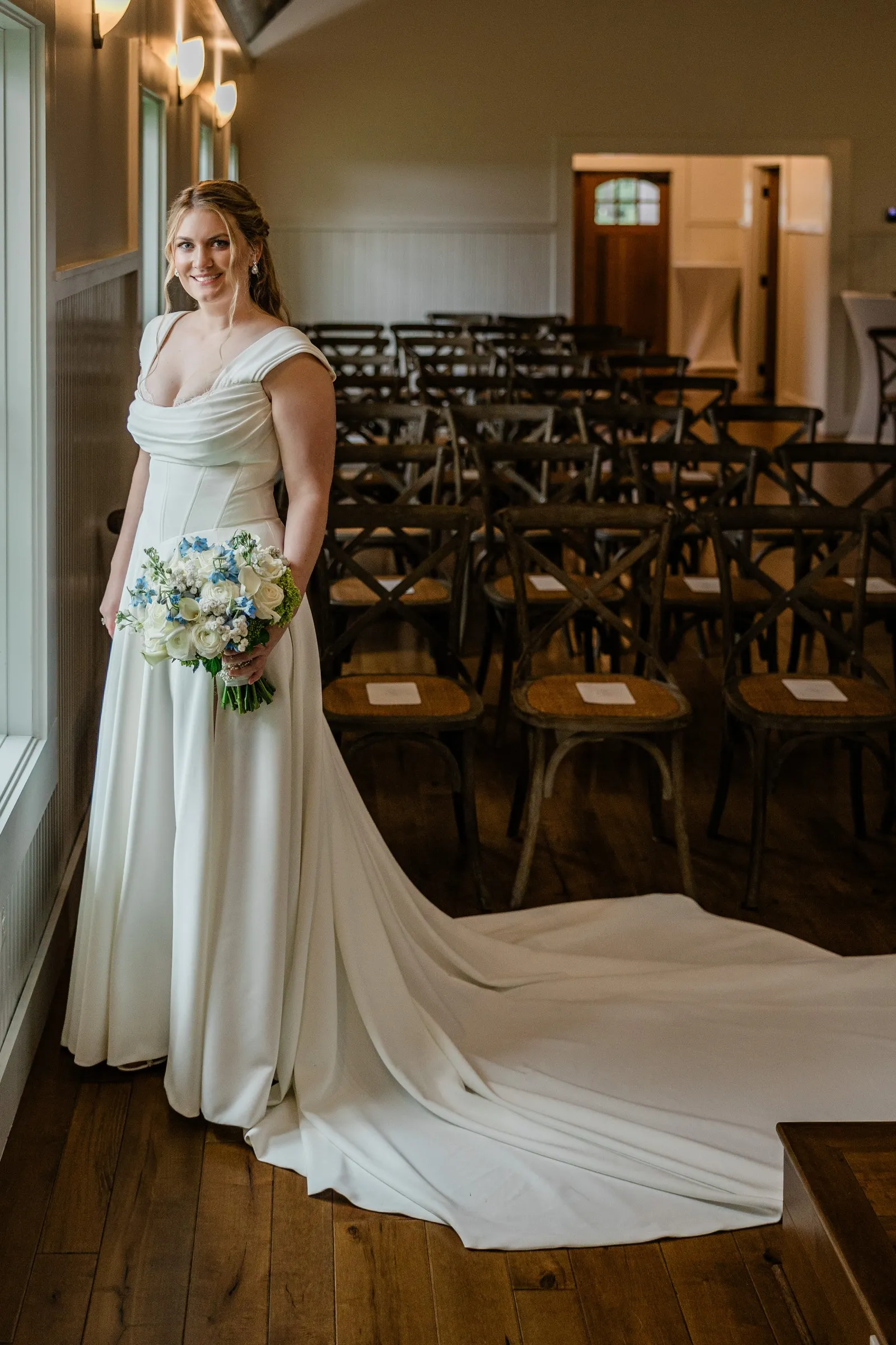 Lauren stands by a Chapel window with her white and blue bouquet and her dress train spread behind her — Tim Larsen Photography, Brainerd Lakes MN