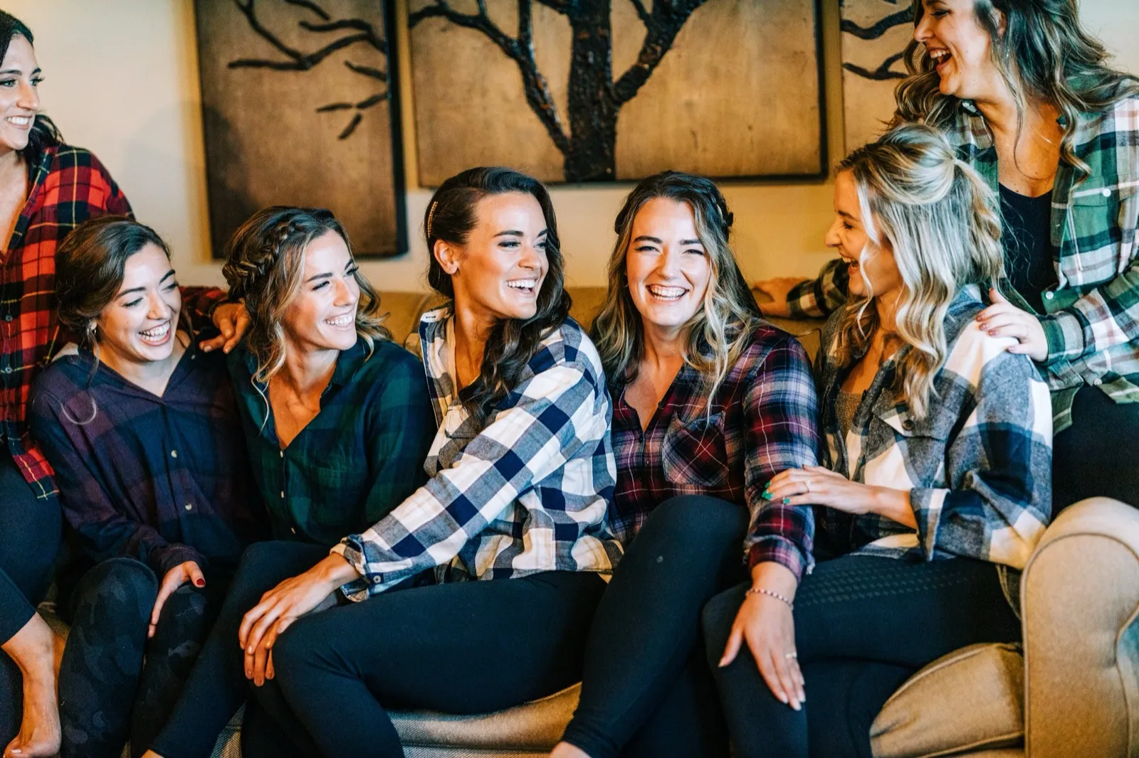 Madison and her bridesmaids in matching flannel shirts laugh together on a couch beneath wood-paneled tree artwork — Tim Larsen Photography, Brainerd Lakes MN