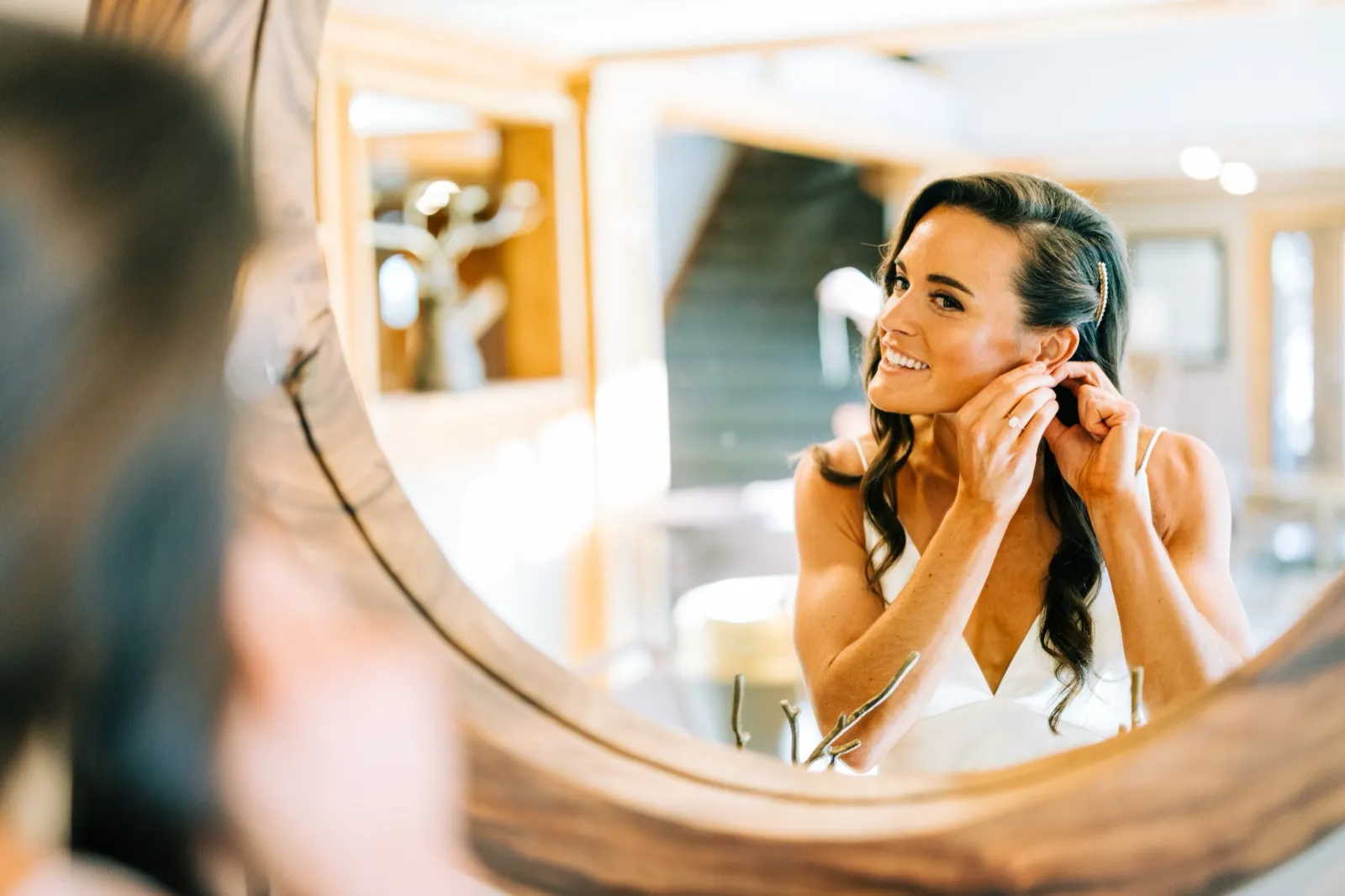 Madison, reflected in a round wood-framed mirror, smiles softly as she fastens an earring in the bridal suite — Tim Larsen Photography, Brainerd Lakes MN