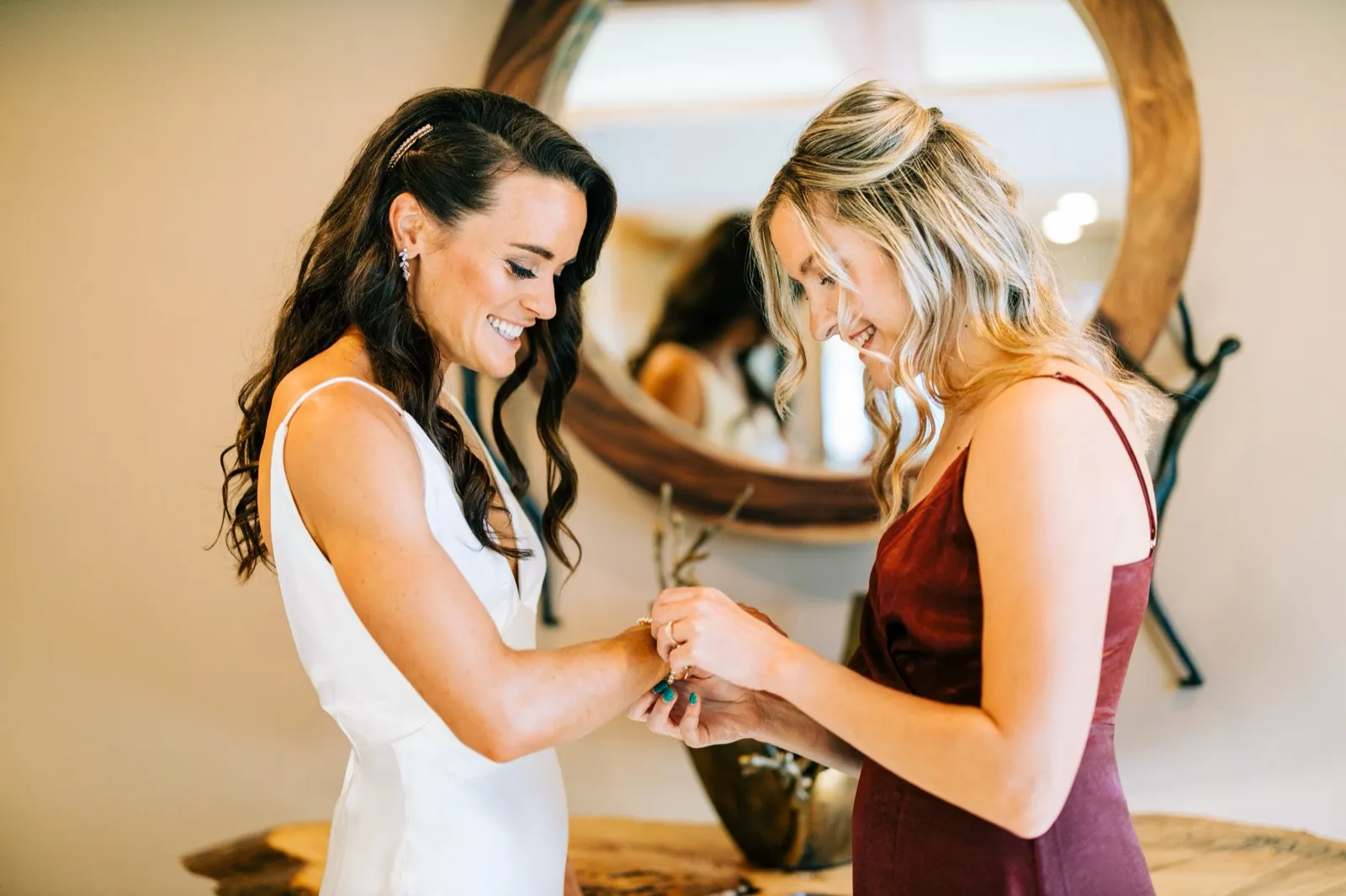 A bridesmaid in burgundy fastens a bracelet on Madison's wrist, a round wood-framed mirror behind them — Tim Larsen Photography, Brainerd Lakes MN