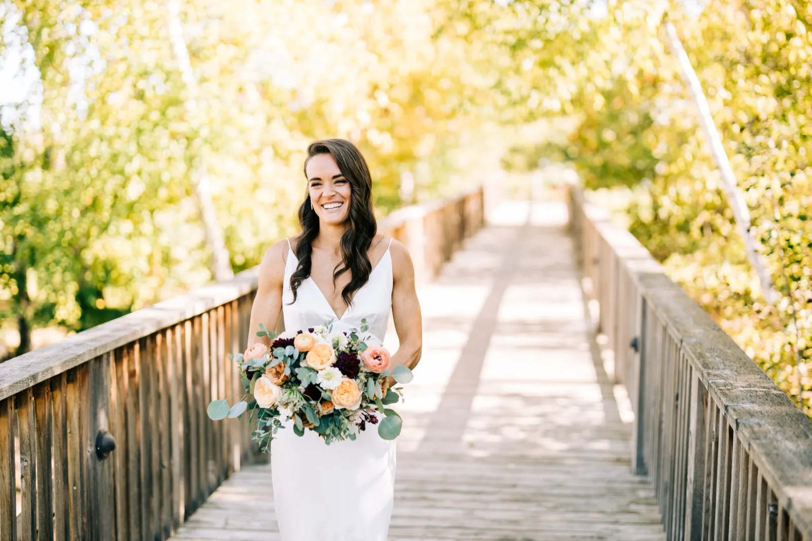 Madison laughs holding her peach-and-burgundy bouquet on a sunlit wooden footbridge framed by autumn trees at Madden's — Tim Larsen Photography, Brainerd Lakes MN