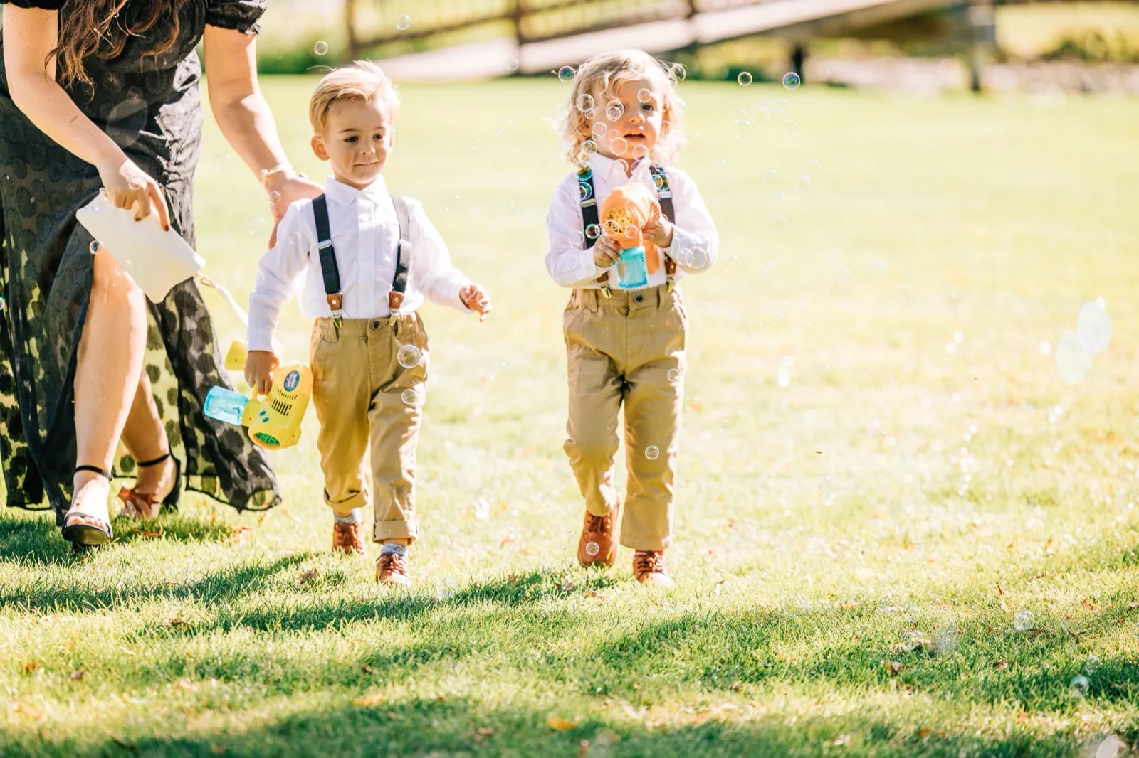 Two young ring bearers in suspenders carry bubble guns across a sunlit lawn as bubbles fill the air before the ceremony — Tim Larsen Photography, Brainerd Lakes MN