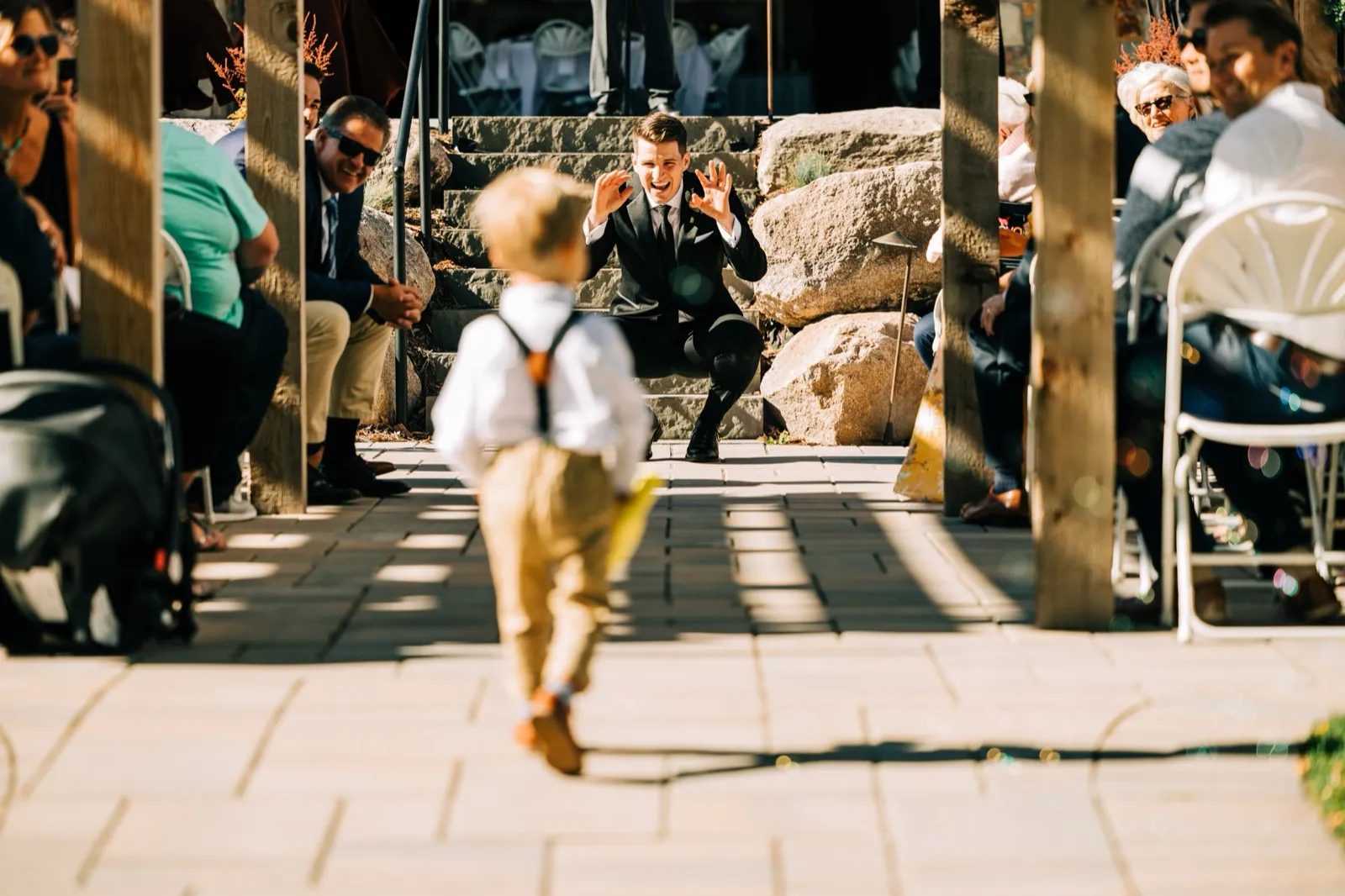 Josh crouches at the end of the aisle, hands raised and grinning, as a small ring bearer walks toward him between the guests — Tim Larsen Photography, Brainerd Lakes MN